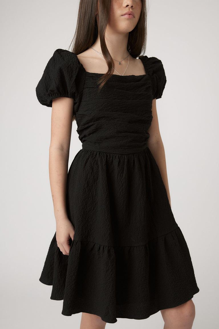 Girl wearing Girls Evie Textured Mini Dress in black with puff sleeves and textured fabric by Decjuba Australian brand