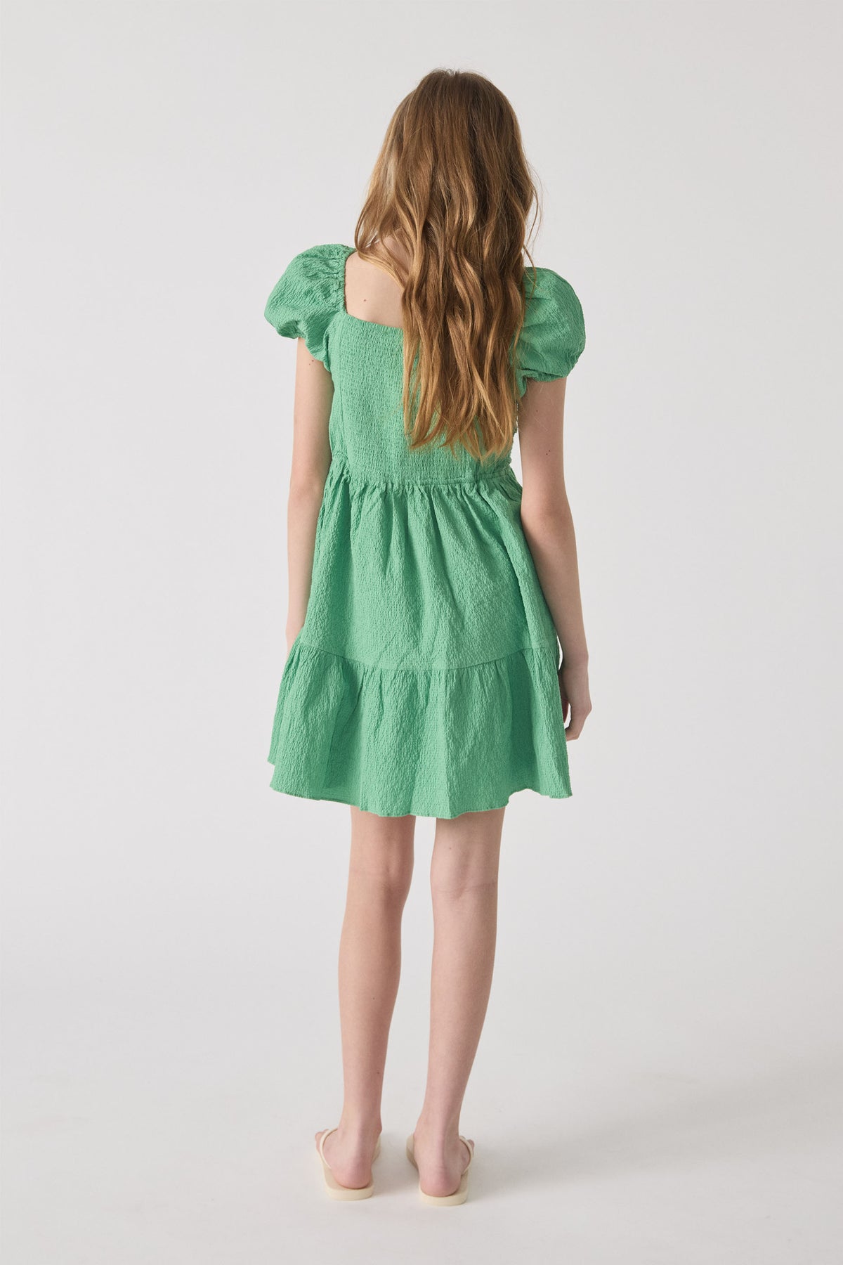 Girl wearing a spring green textured mini dress with puff sleeves and tiered skirt, shown from the back view.