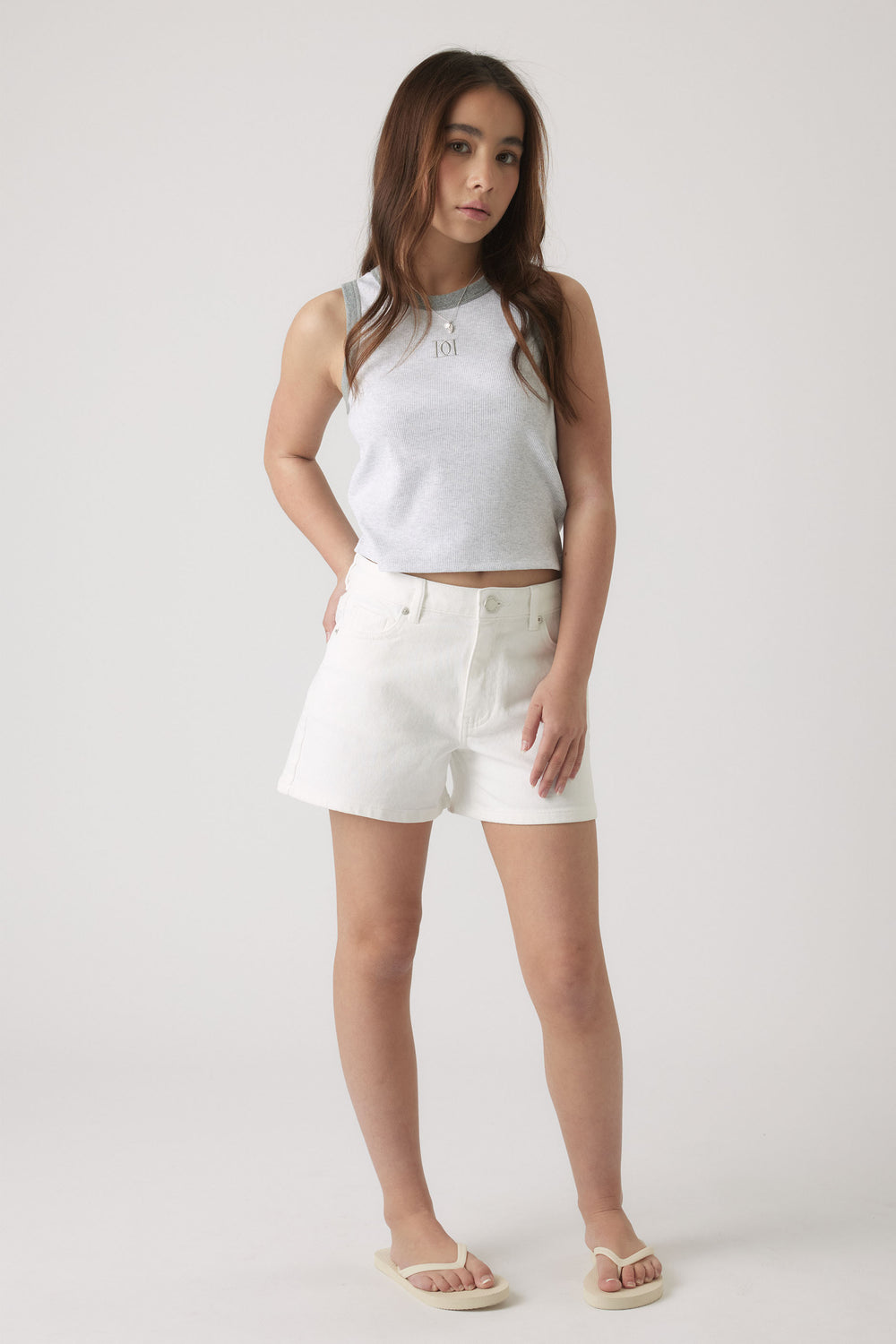 Teen girl wearing a light grey sleeveless tank paired with white high-waisted shorts and beige sandals, standing confidently.