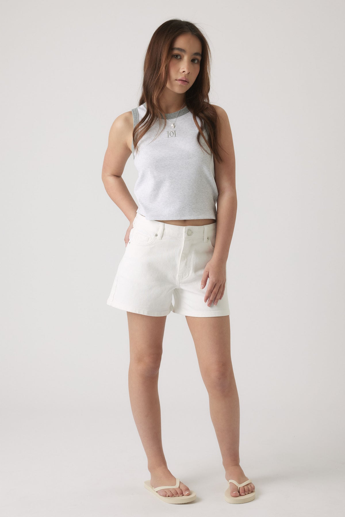 Teen girl wearing a light grey sleeveless tank paired with white high-waisted shorts and beige sandals, standing confidently.