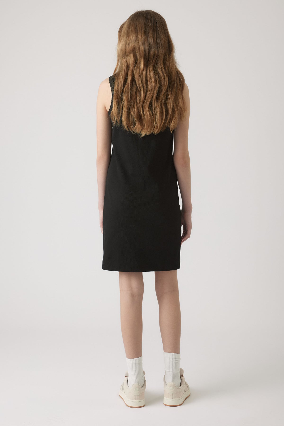 Girl wearing a simple sleeveless black dress paired with white socks and casual off-white sneakers against a plain background