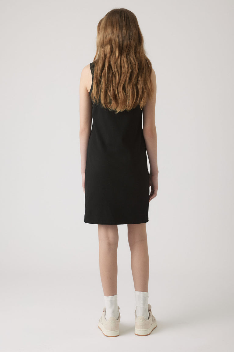 Girl wearing a simple sleeveless black dress paired with white socks and casual off-white sneakers against a plain background