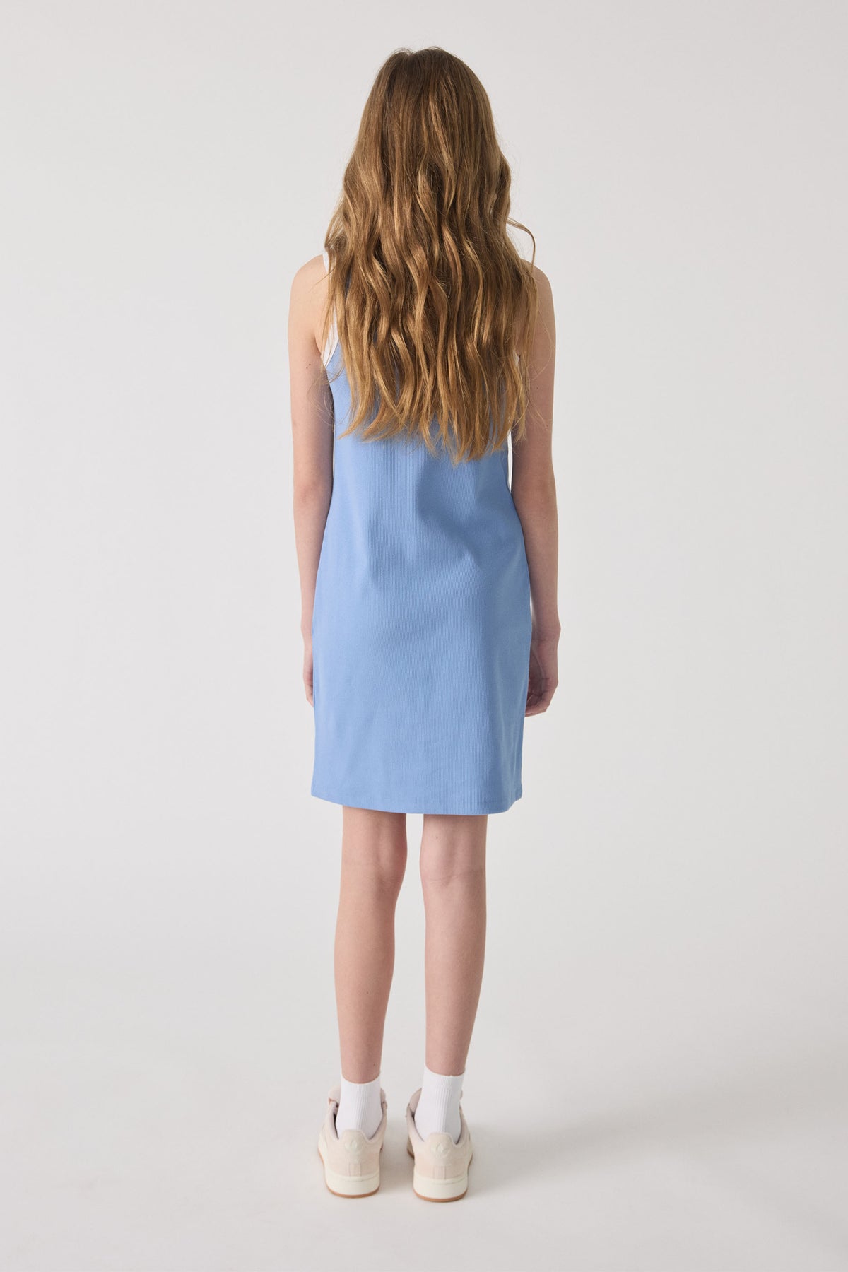 Girl wearing a sleeveless dusty blue monogram dress paired with white socks and casual sneakers, viewed from the back.