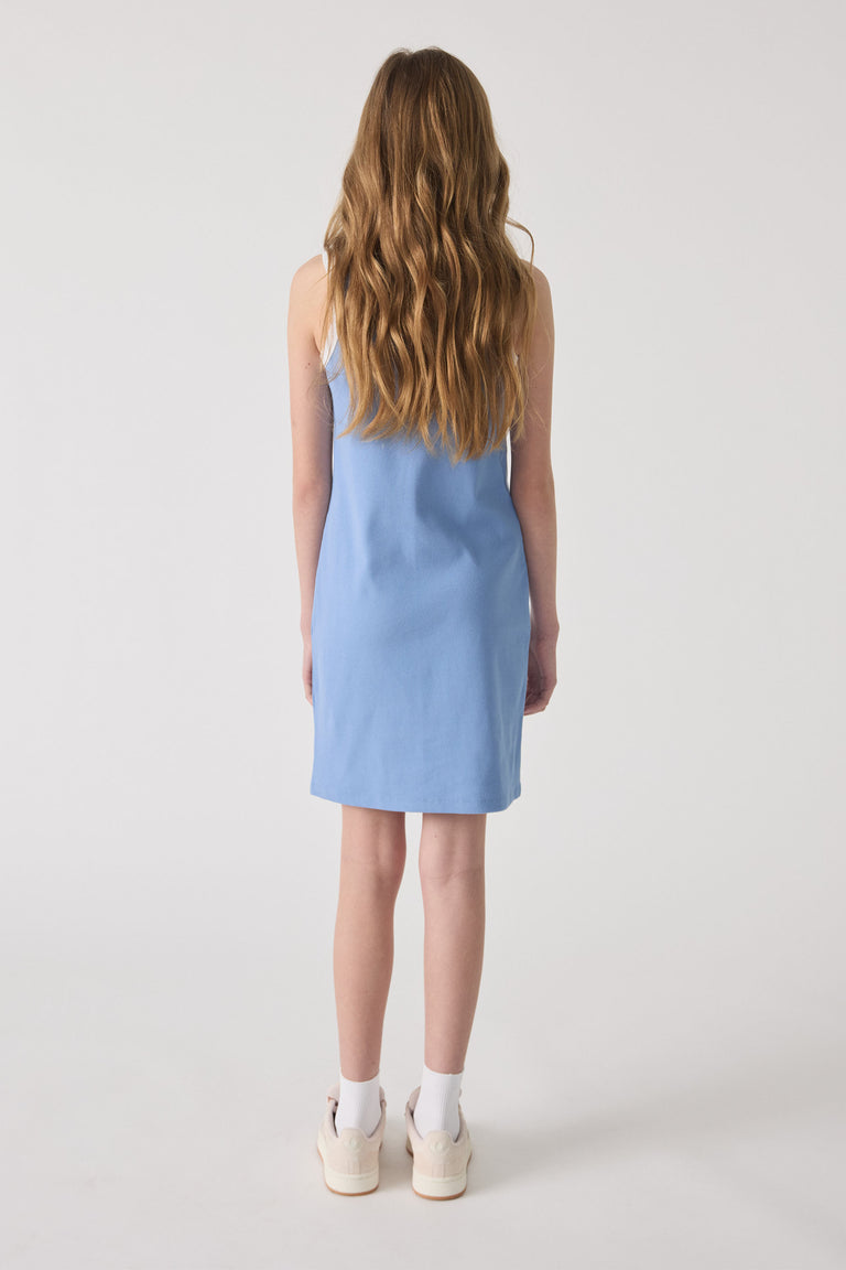 Girl wearing a sleeveless dusty blue monogram dress paired with white socks and casual sneakers, viewed from the back.