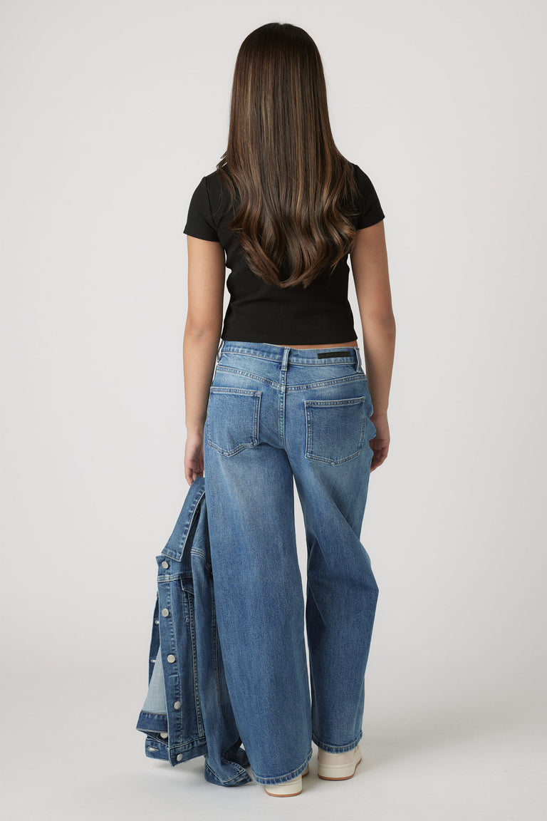 Teen girl seen from behind wearing a black crop tee and wide-leg blue jeans holding a denim jacket casually.
