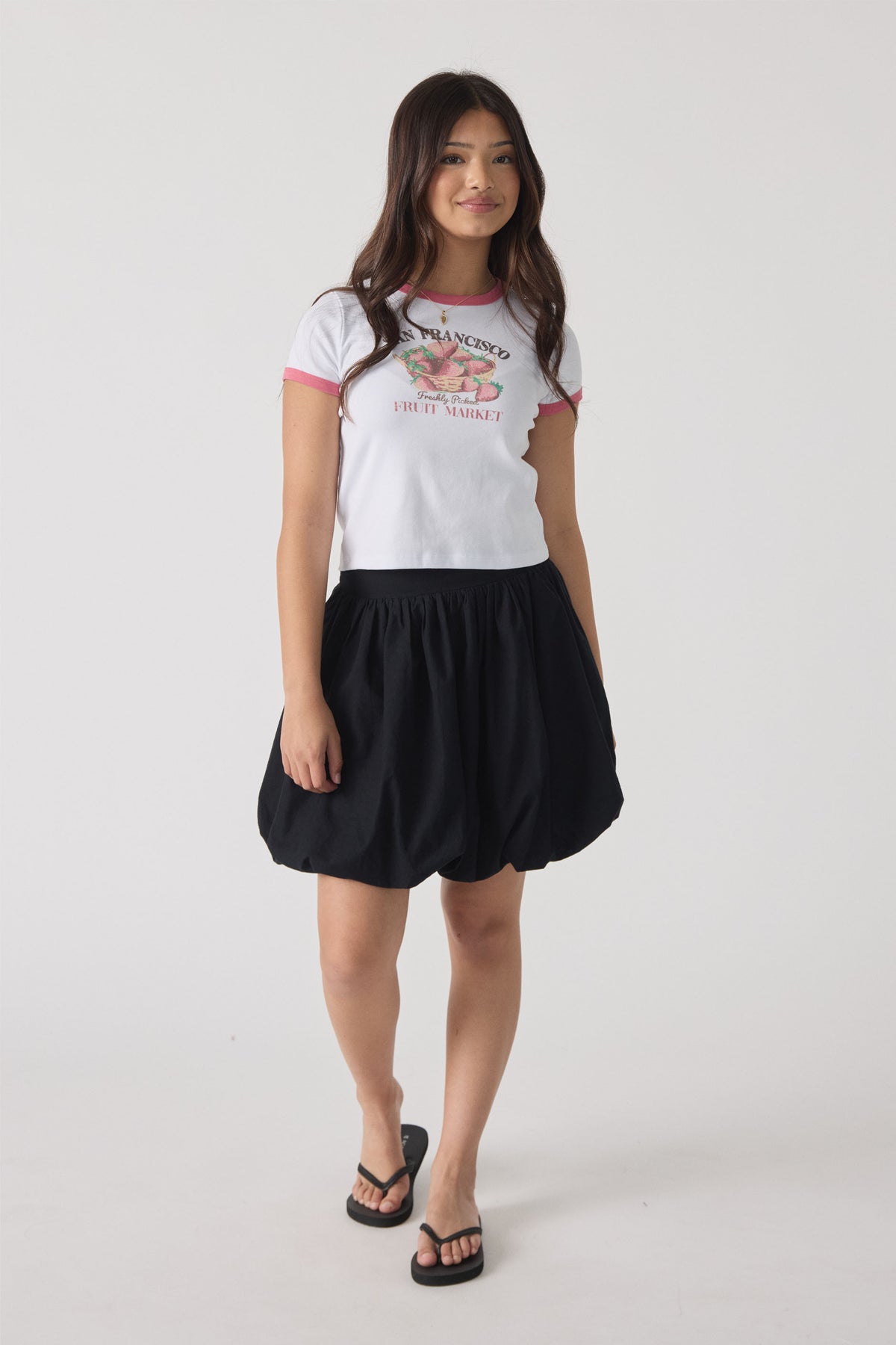 Girl wearing a black bubble mini skirt paired with a white graphic tee and black sandals, smiling at the camera.