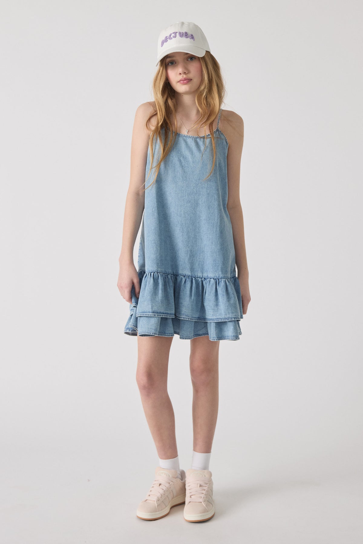 Teen girl wearing light denim tiered dress paired with white embroidered cap and pale pink sneakers against plain background