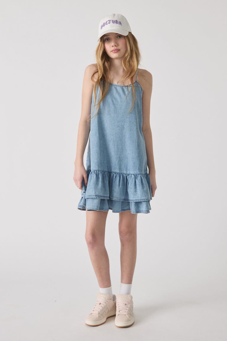 Teen girl wearing light denim tiered dress paired with white embroidered cap and pale pink sneakers against plain background