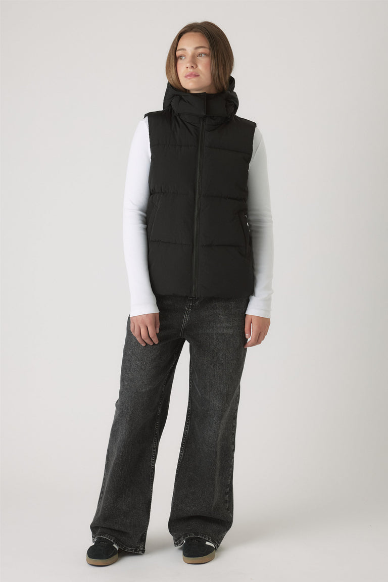 Girls Hooded Puffer Vest in black with a quilted design, high collar, and zip closure styled over white long sleeves.