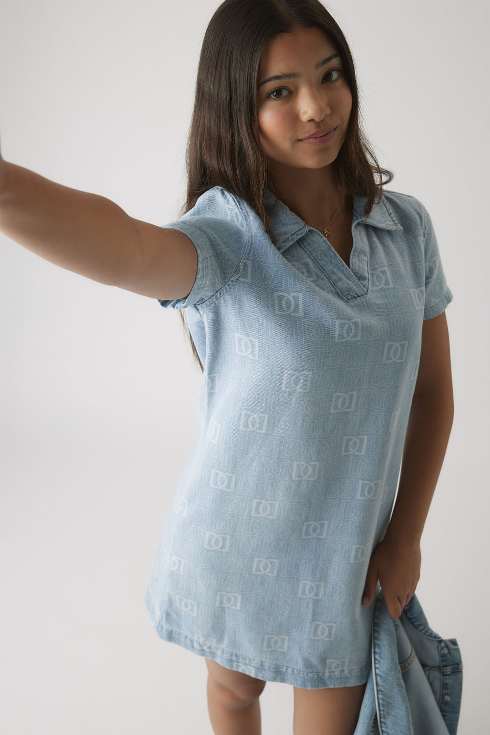 Girl wearing a light blue denim dress with subtle monogram pattern and a collar, holding a matching denim jacket.