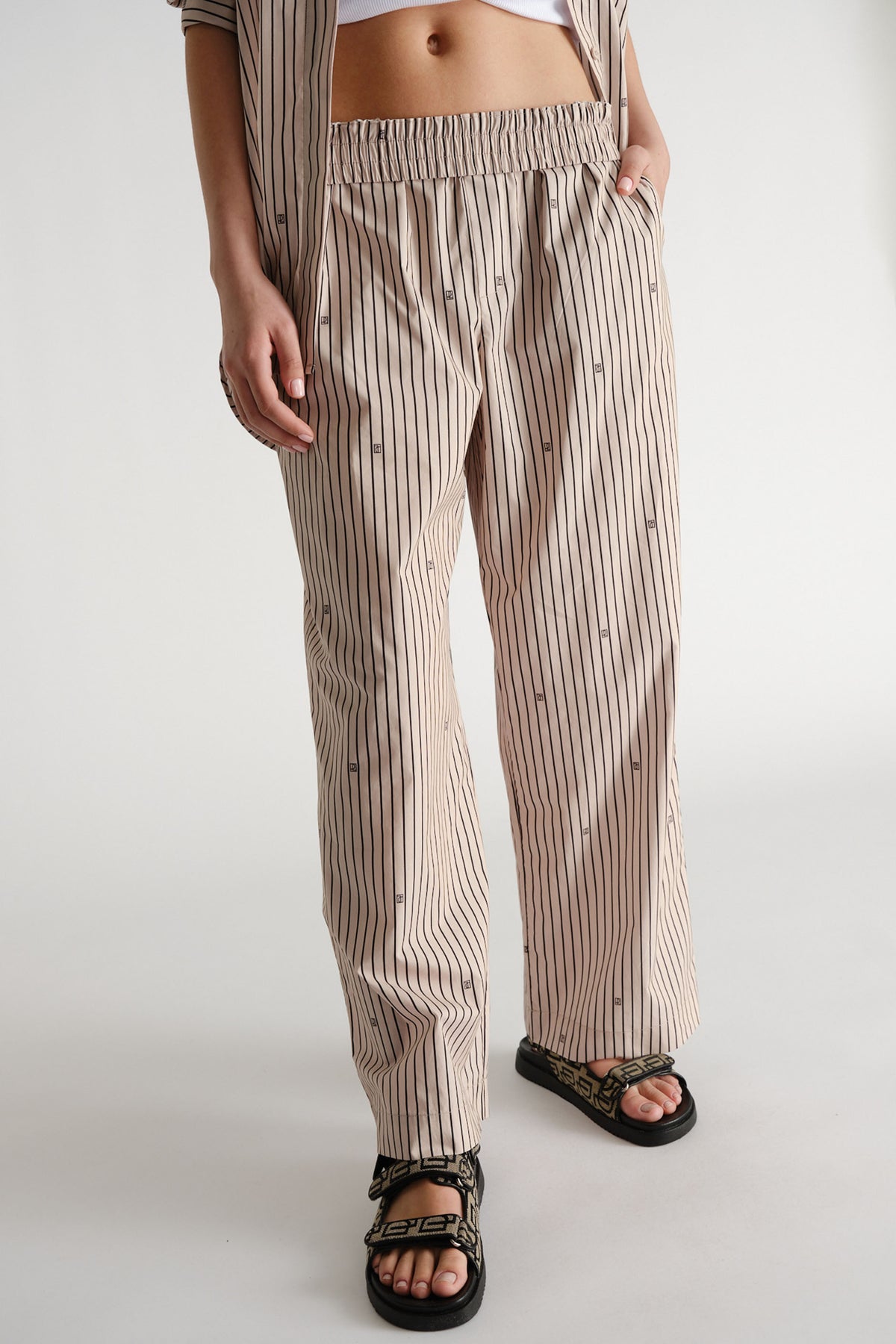 Relaxed fit beige pants with vertical monogram stripes, elastic waist, paired with black patterned sandals.