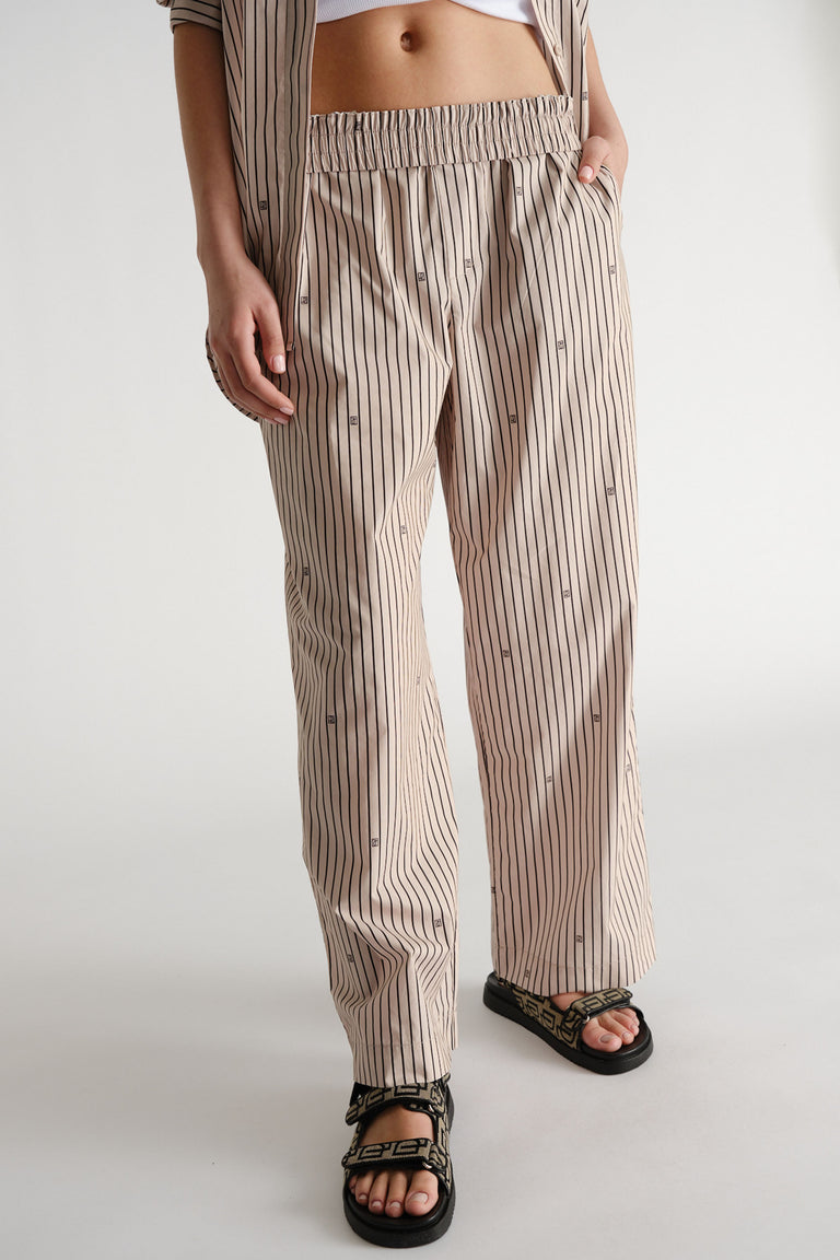 Relaxed fit beige pants with vertical monogram stripes, elastic waist, paired with black patterned sandals.