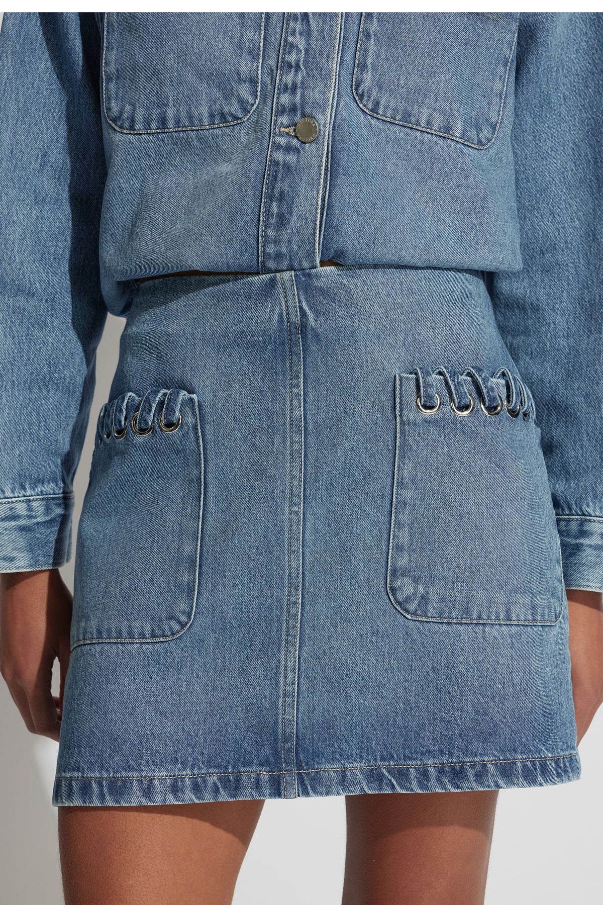 Close-up of a mid-blue denim skirt with front pockets featuring silver eyelet detailing for a modern, casual look.