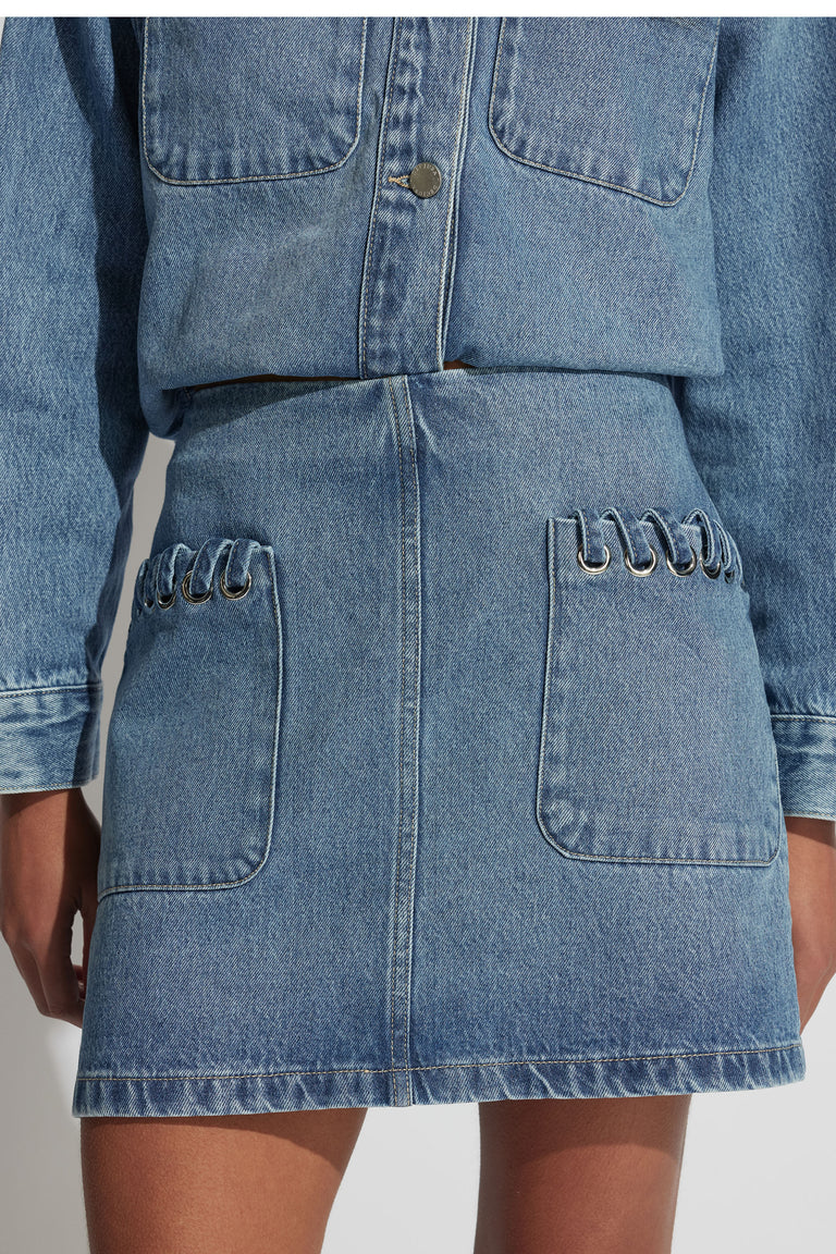 Close-up of a mid-blue denim skirt with front pockets featuring silver eyelet detailing for a modern, casual look.