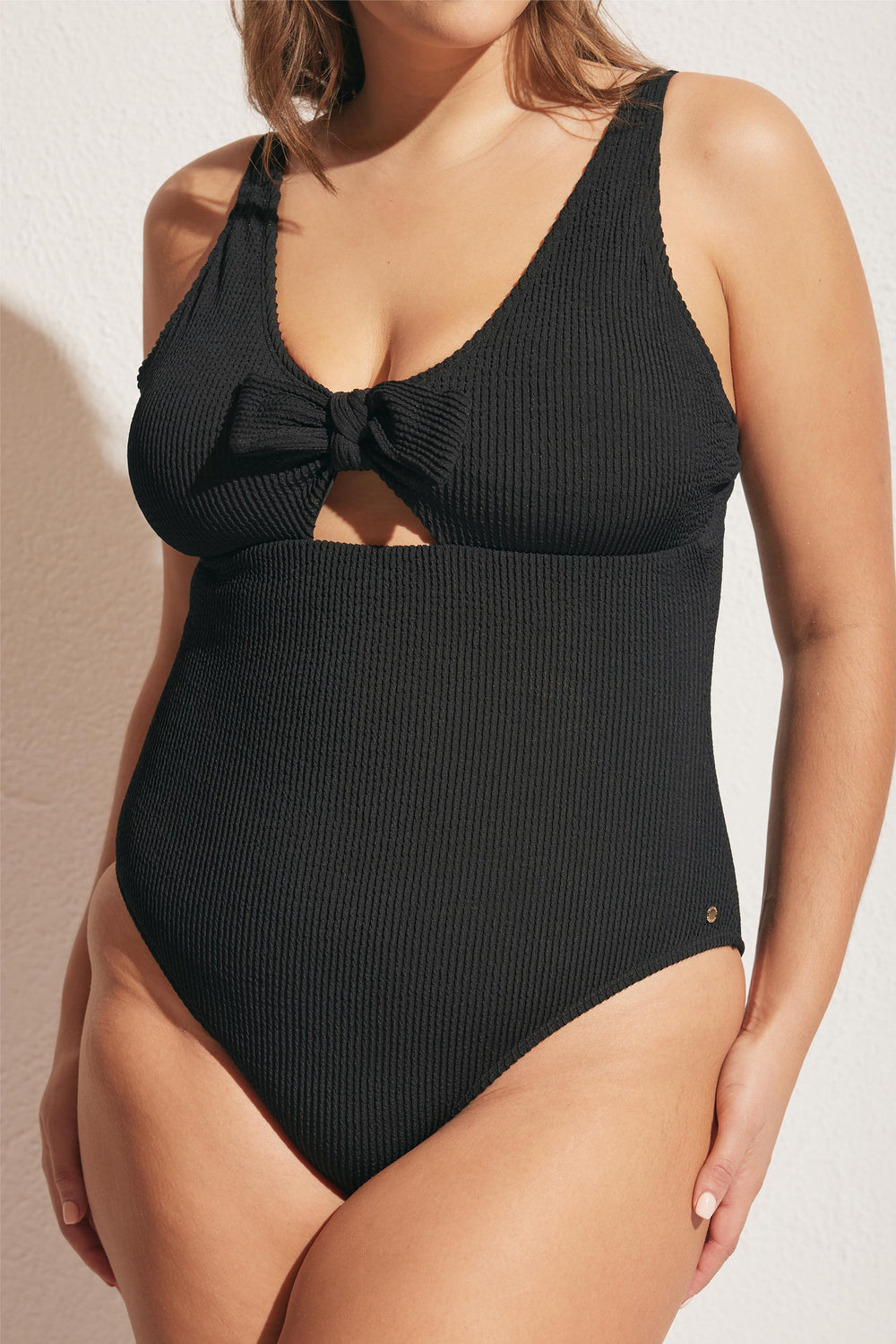 Woman wearing a textured black one-piece swimsuit with a front tie knot and subtle cutout detail beneath the bust