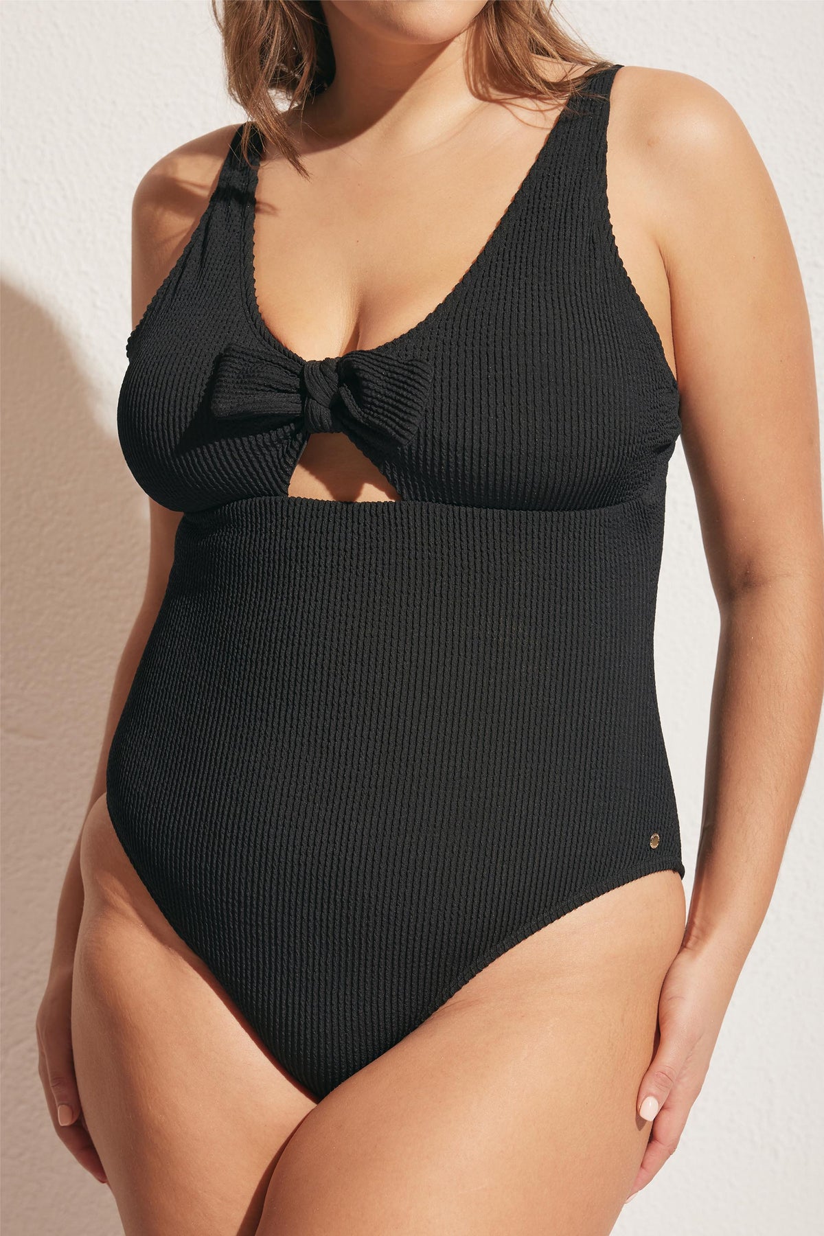 Woman wearing a textured black one-piece swimsuit with a front tie knot and subtle cutout detail beneath the bust