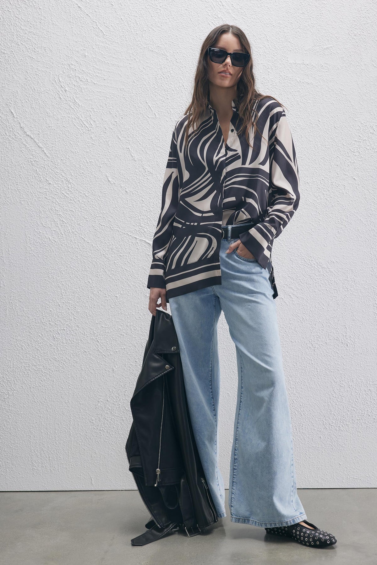 Woman wearing loose light blue wide-leg jeans with a graphic black and beige shirt, holding a black jacket for a chic look.