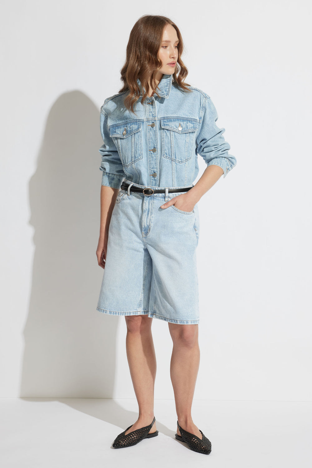 Woman wearing light blue longline denim shorts with a matching cropped jacket and black belt, paired with black flats.