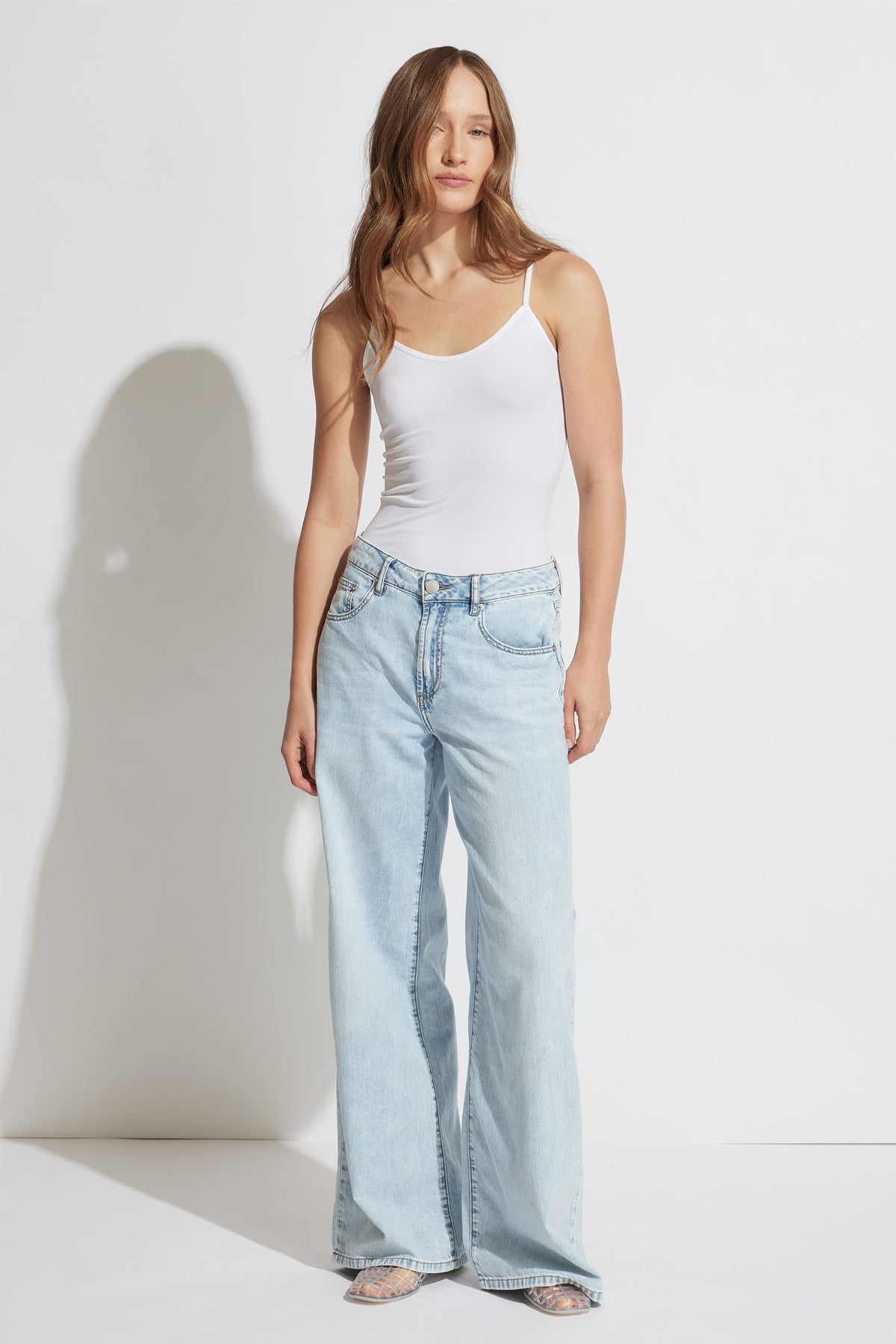Woman wearing a fitted white thin strap tank top paired with relaxed light-wash wide-leg denim jeans and clear heels