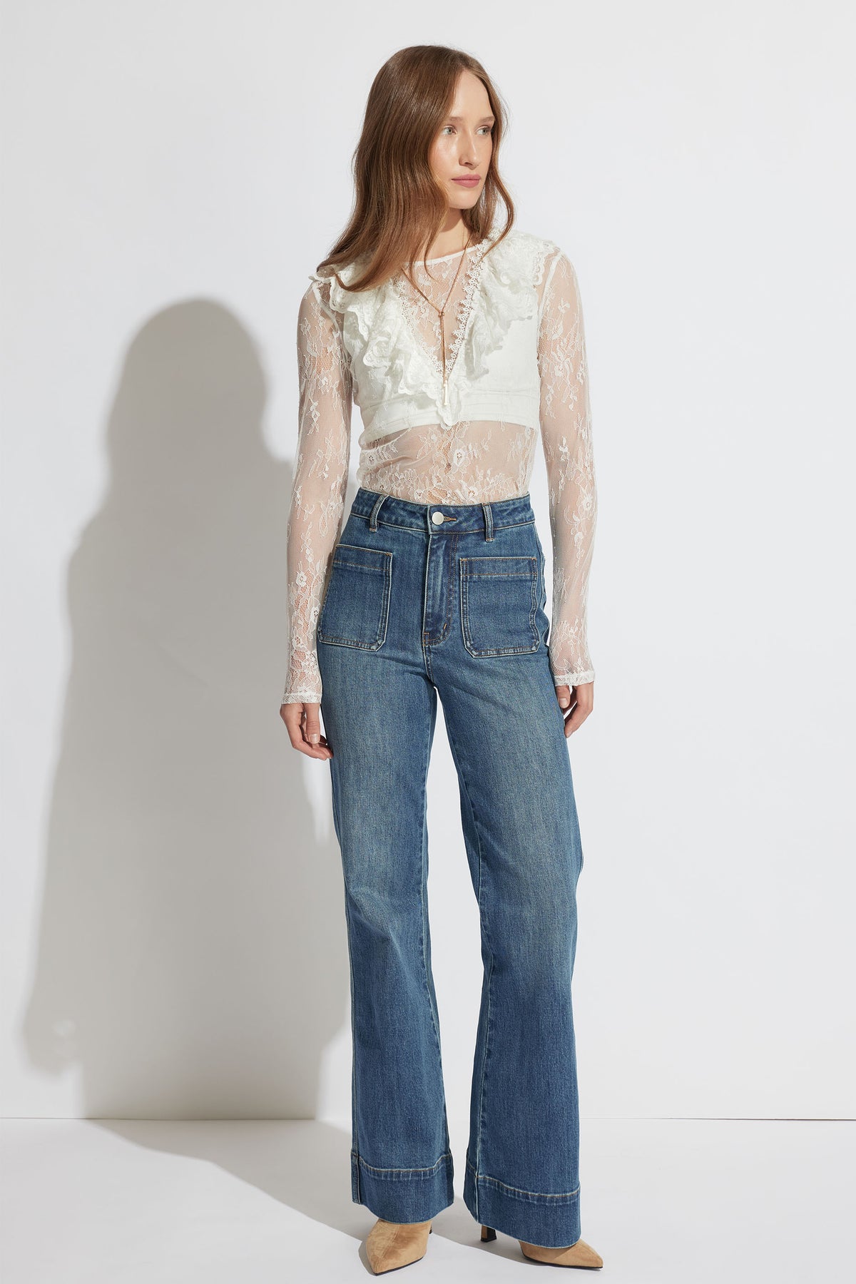 Woman wearing a sheer lace white top with ruffle detail paired with high-waisted wide-leg blue jeans and tan shoes