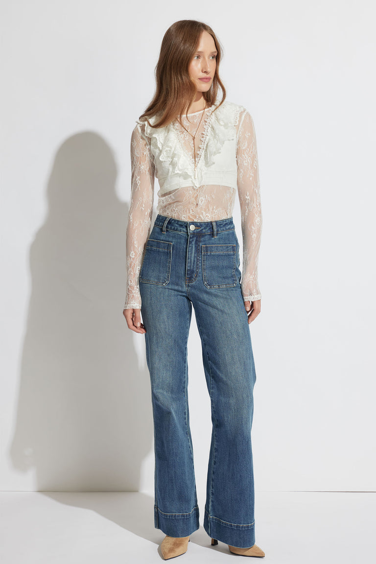 Woman wearing a sheer lace white top with ruffle detail paired with high-waisted wide-leg blue jeans and tan shoes