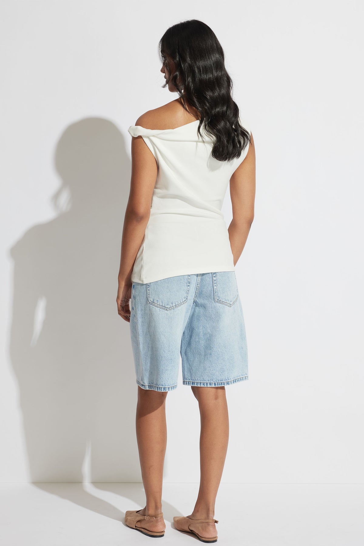 Back view of model wearing a white twist shoulder top with an asymmetric hem and light wash denim shorts