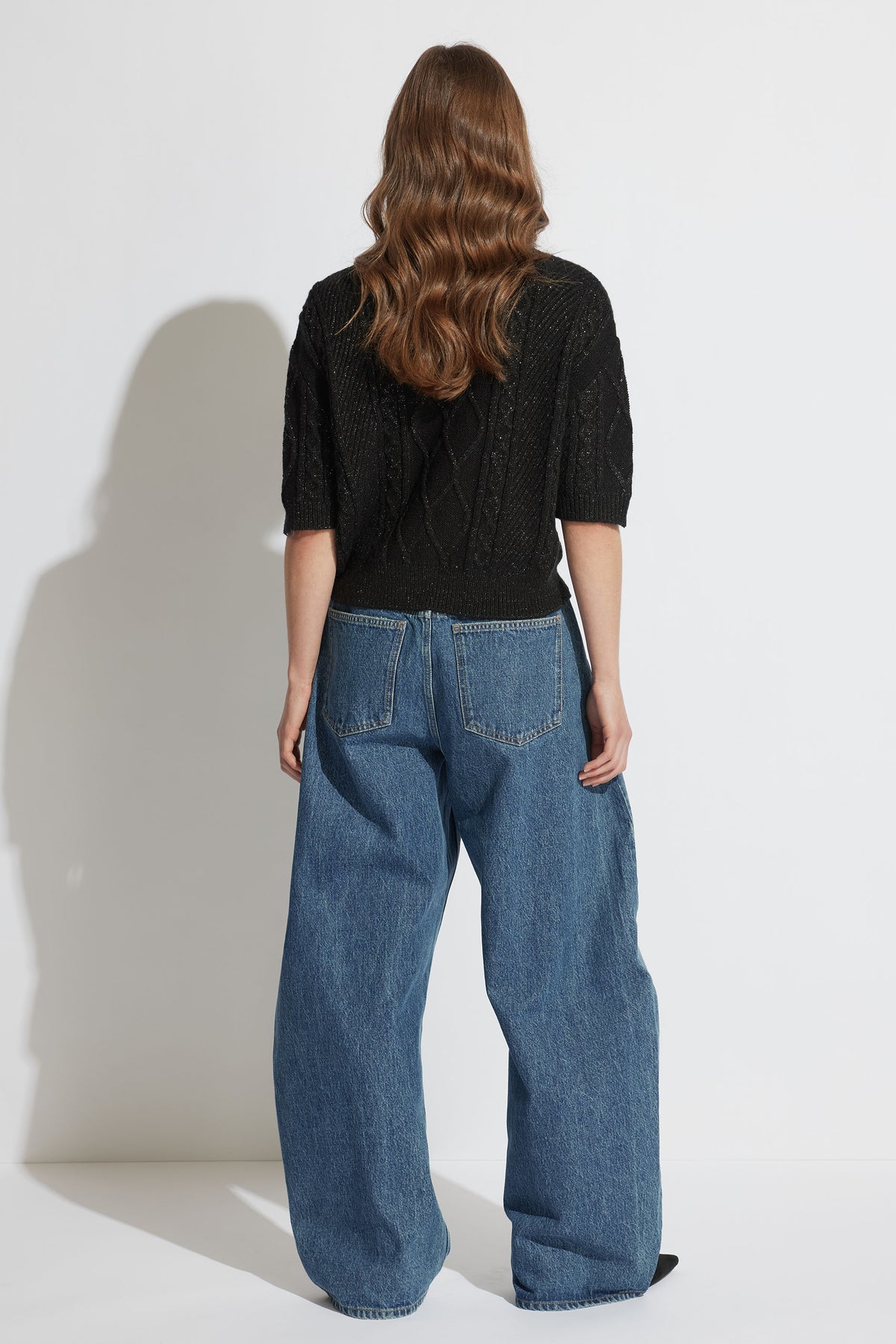 Back view of model wearing Sasha Lurex Cable Knit Jumper with detachable sleeves and loose denim jeans, portrait format.