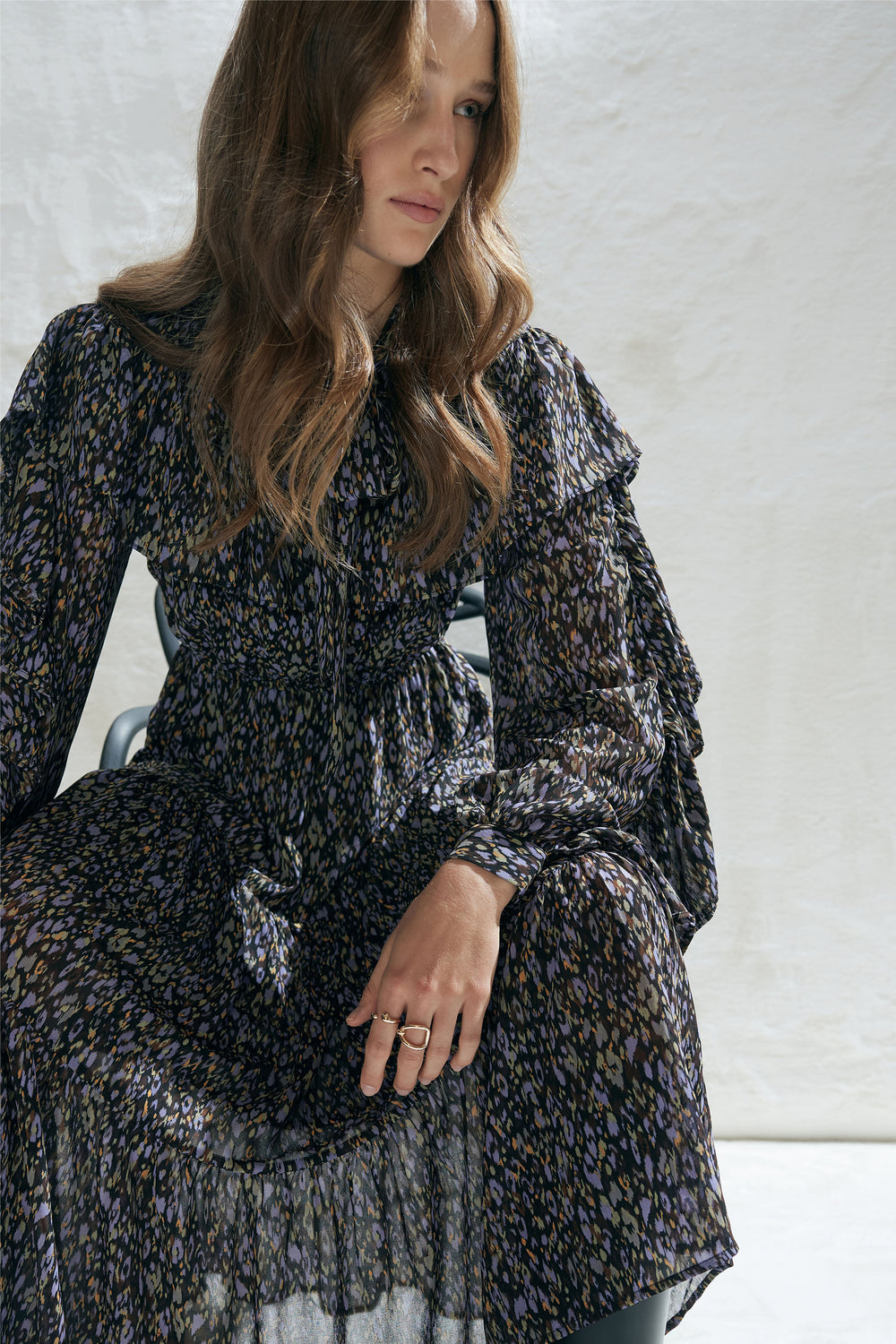 Woman wearing a patterned midi dress with ruffle details and long sleeves sitting against a neutral background.
