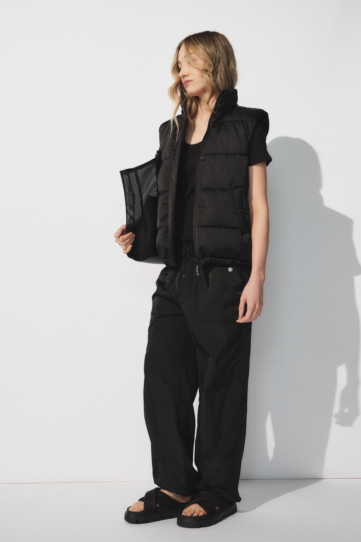 Woman wearing a black sleeveless puffer vest with black relaxed joggers and black sandals against a white background