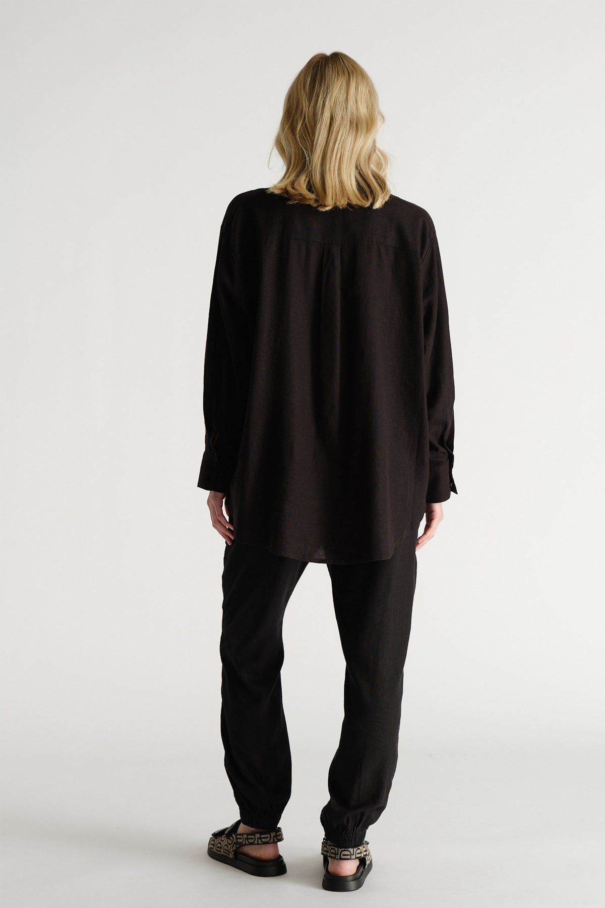 Back view of a woman wearing relaxed black linen-blend joggers paired with a loose black long-sleeve shirt and patterned sandals.