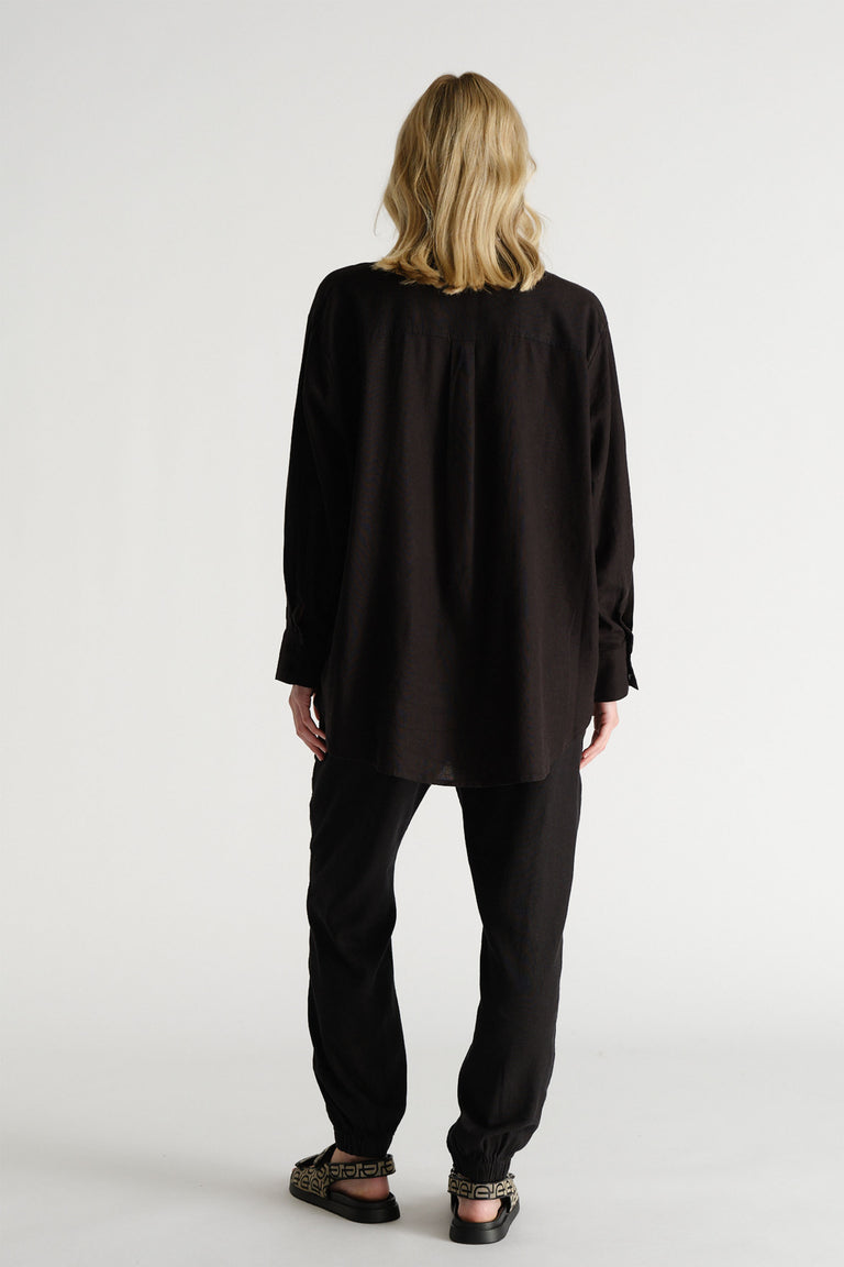 Back view of a woman wearing relaxed black linen-blend joggers paired with a loose black long-sleeve shirt and patterned sandals.
