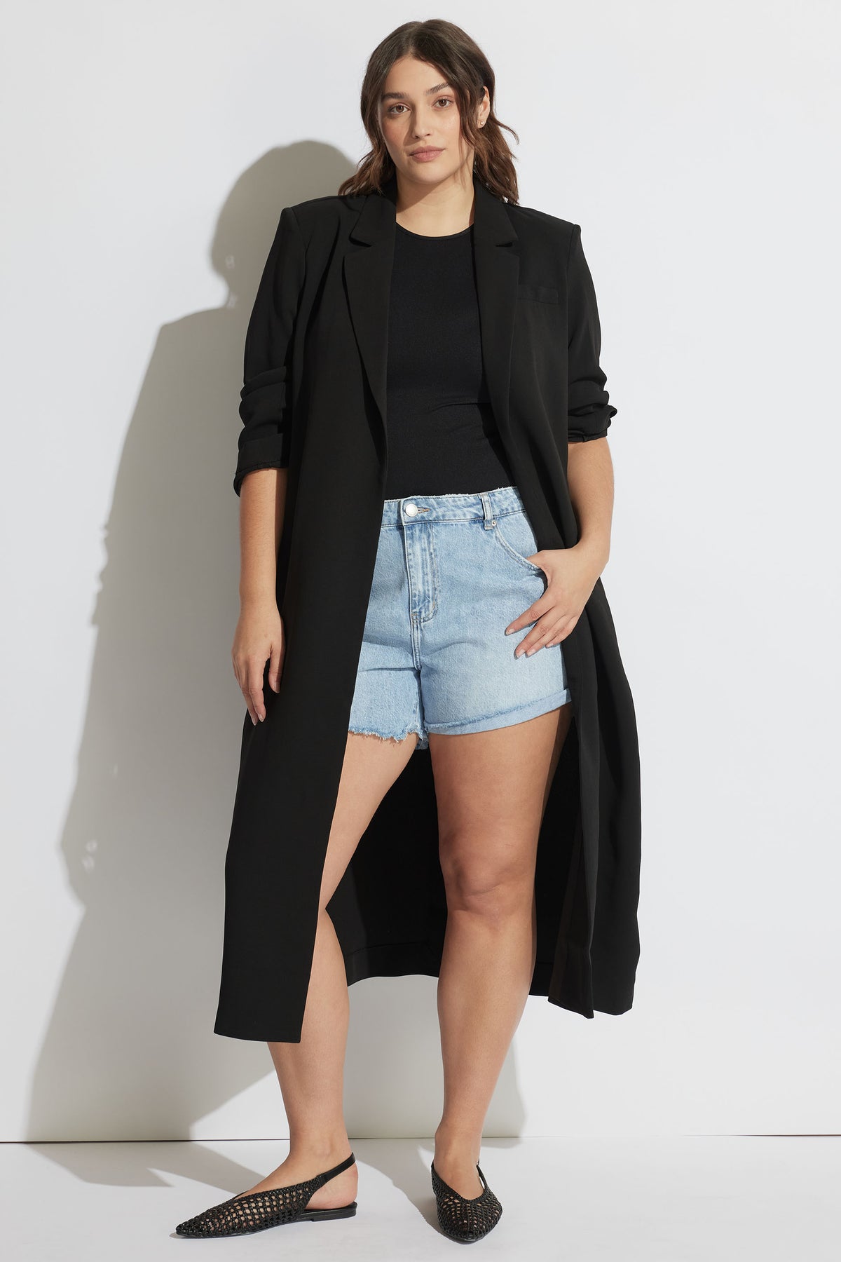 Woman wearing light blue rolled cuff denim shorts with a black longline blazer, black top, and woven flats.