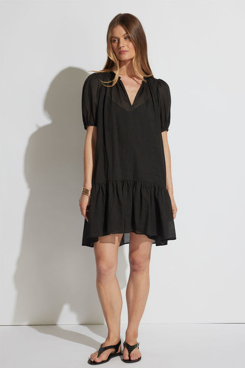 Woman wearing a loose black mini dress with puff sleeves and a ruffled hem paired with black sandals against a white background.