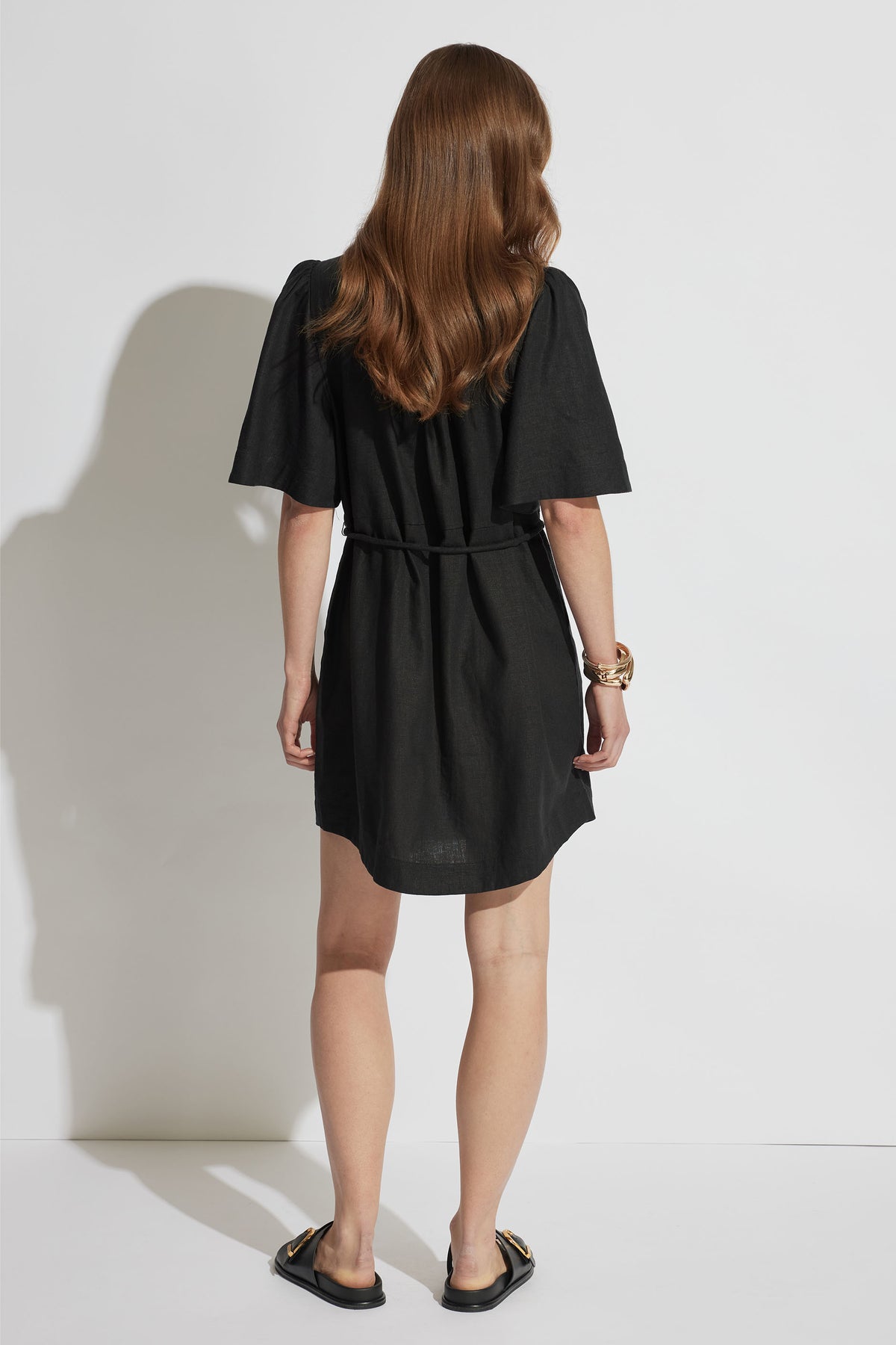 Woman wearing a black mini shirt dress with flutter sleeves and a waist tie, paired with black slip-on sandals, shown from behind.