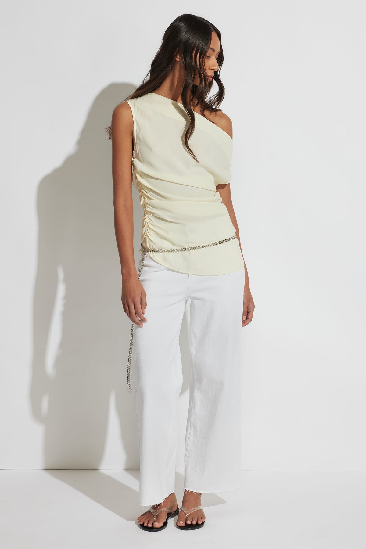 Woman wearing a pale yellow one-shoulder ruched top with white wide-leg pants and sandals, accessorized with a chain belt