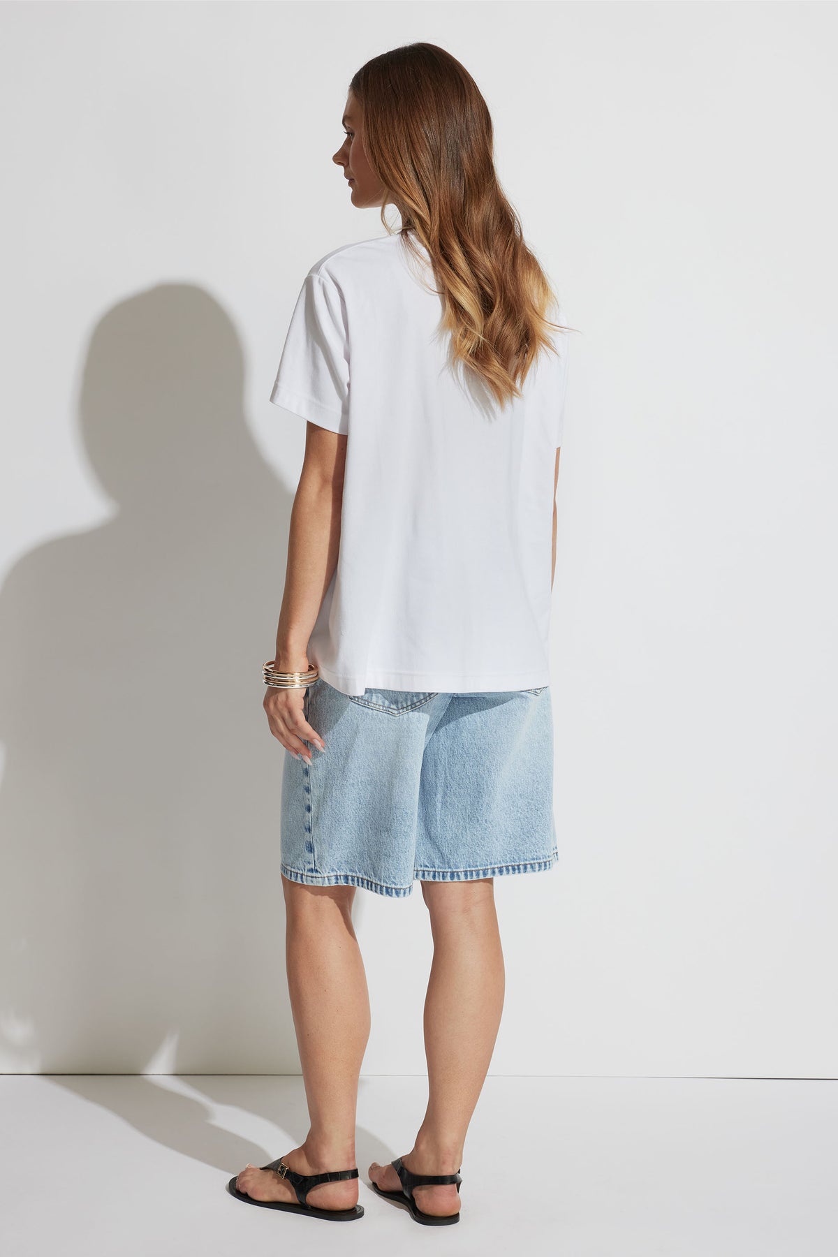 Woman wearing a relaxed white tee paired with light wash loose denim shorts and black sandals, viewed from behind.