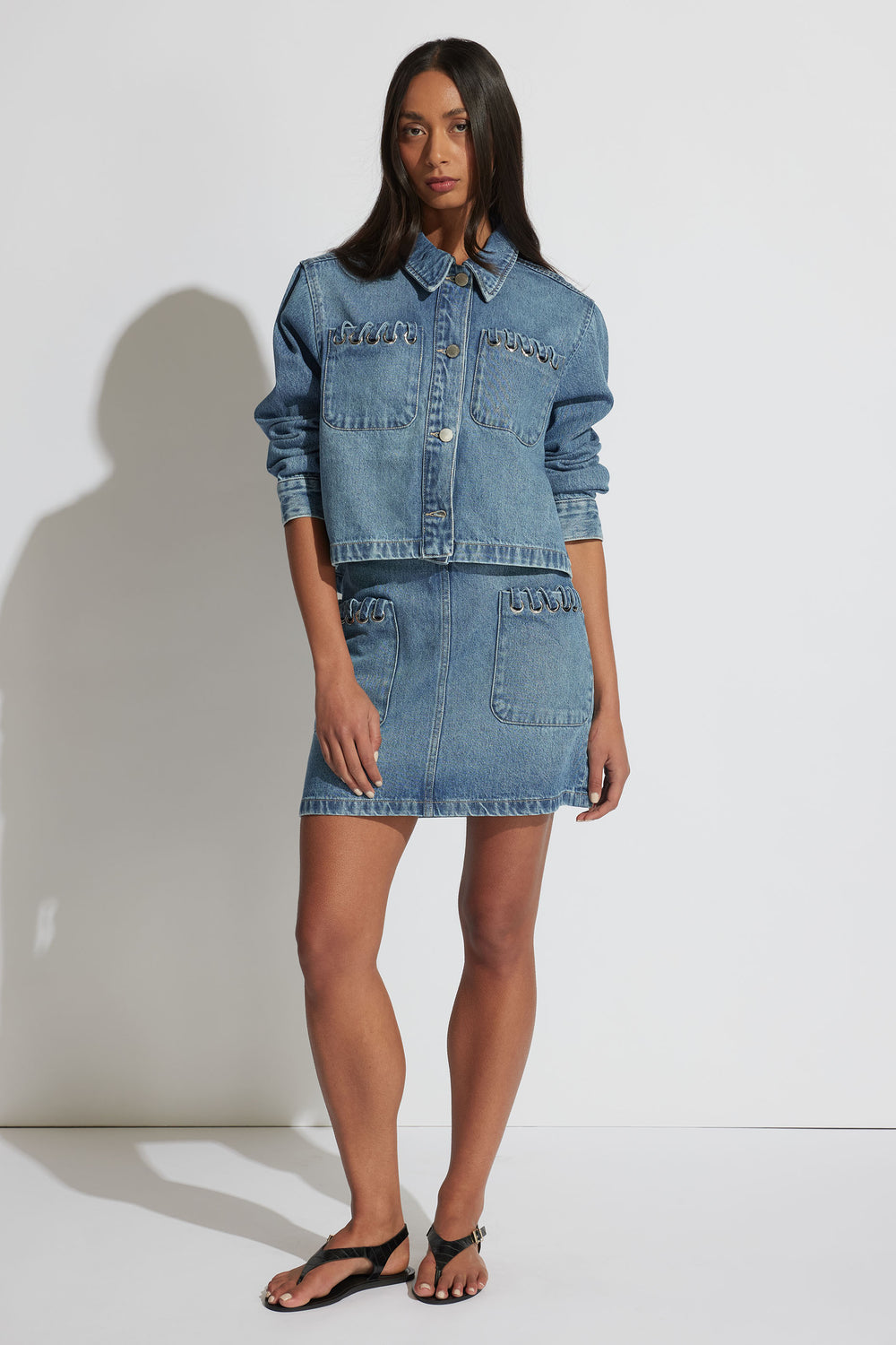 Woman wearing a medium blue denim jacket with front pockets paired with a matching denim mini skirt and black sandals