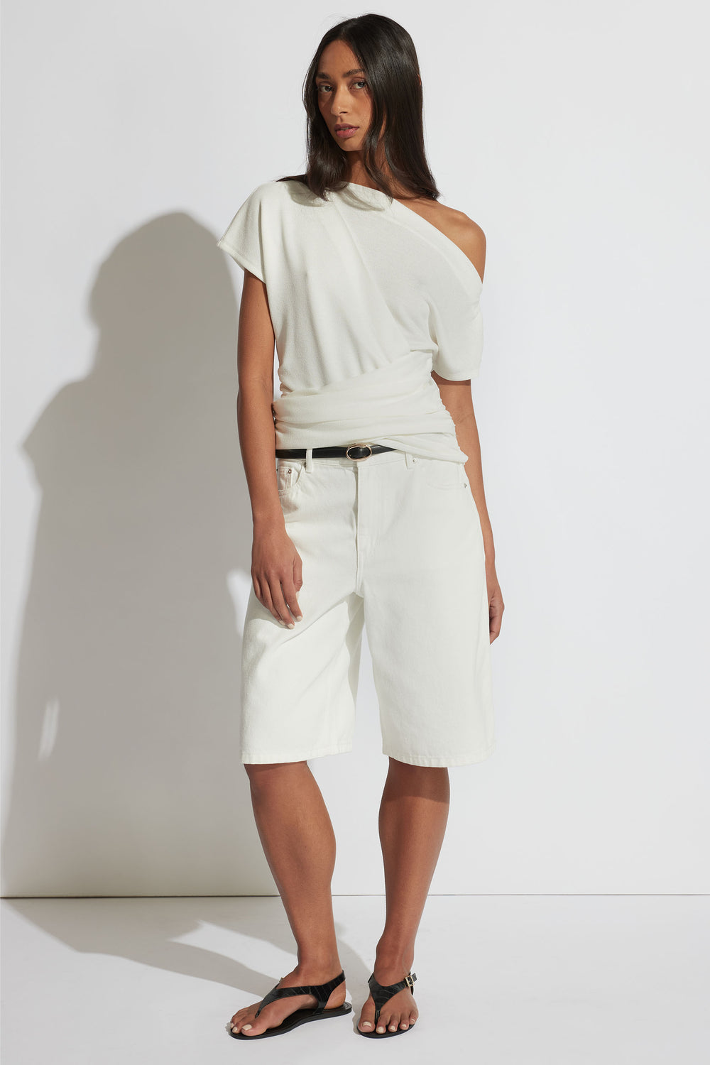 Woman wearing an off-shoulder gathered ivory top paired with loose white bermuda shorts and black sandals.