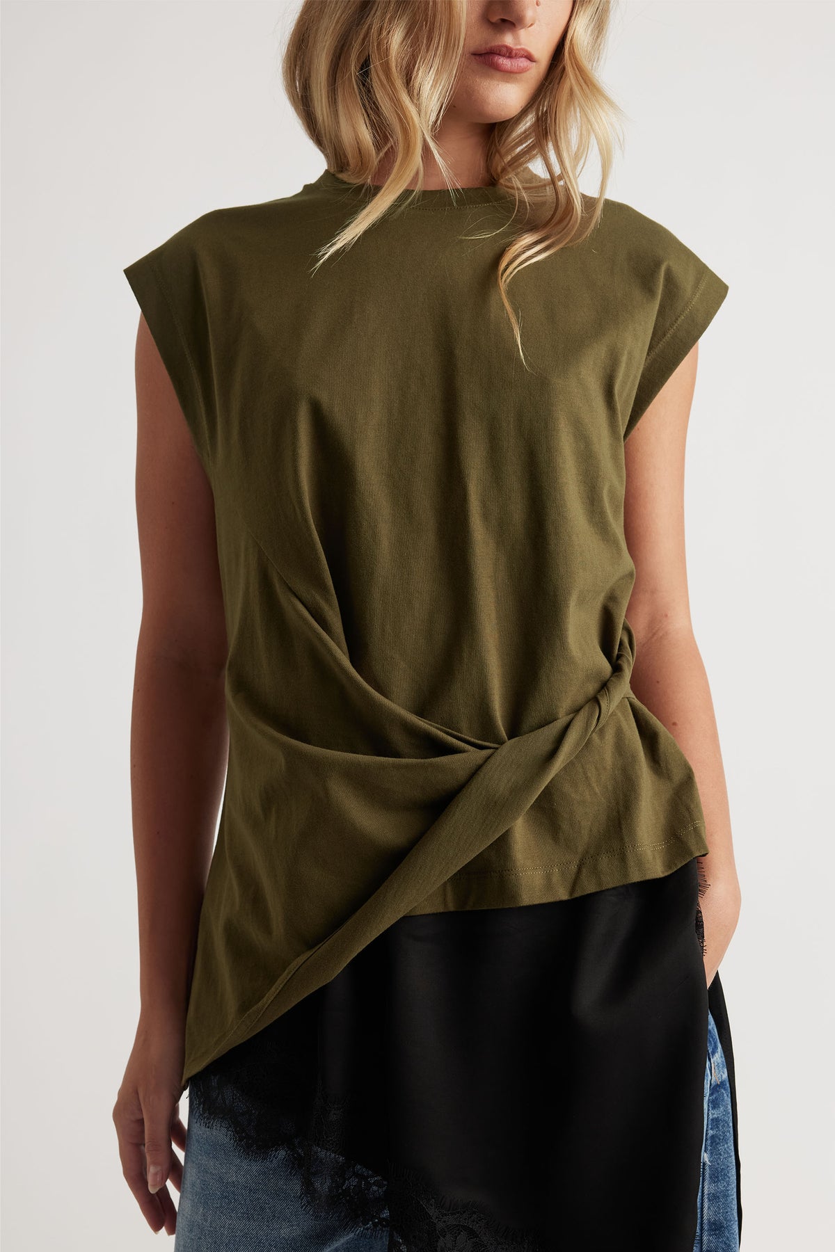 Woman wearing an olive green top with a twisted detail at the waist, paired with layered black and denim bottoms.
