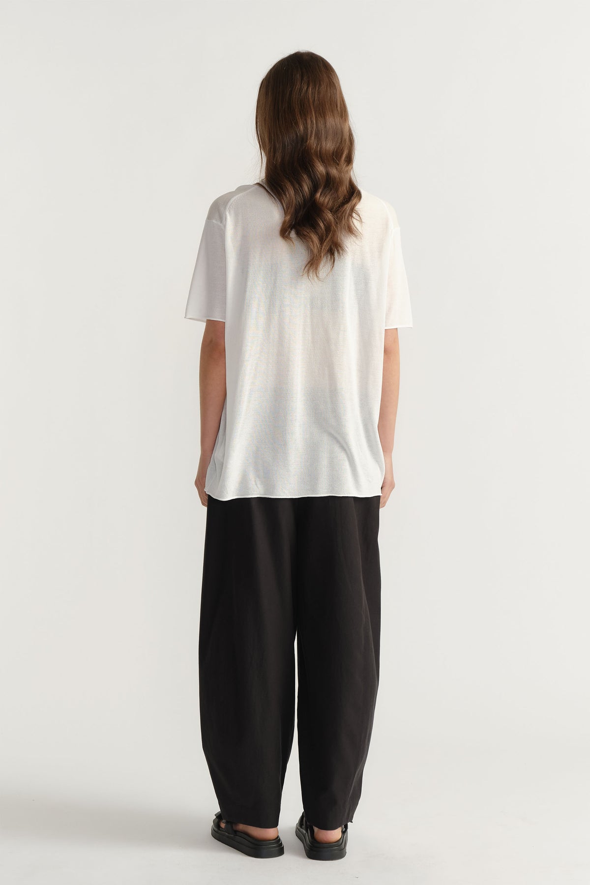 Back view portrait of model wearing white D-Luxe Fine Knit Tee with short sleeves and black pants in gallery position 3.