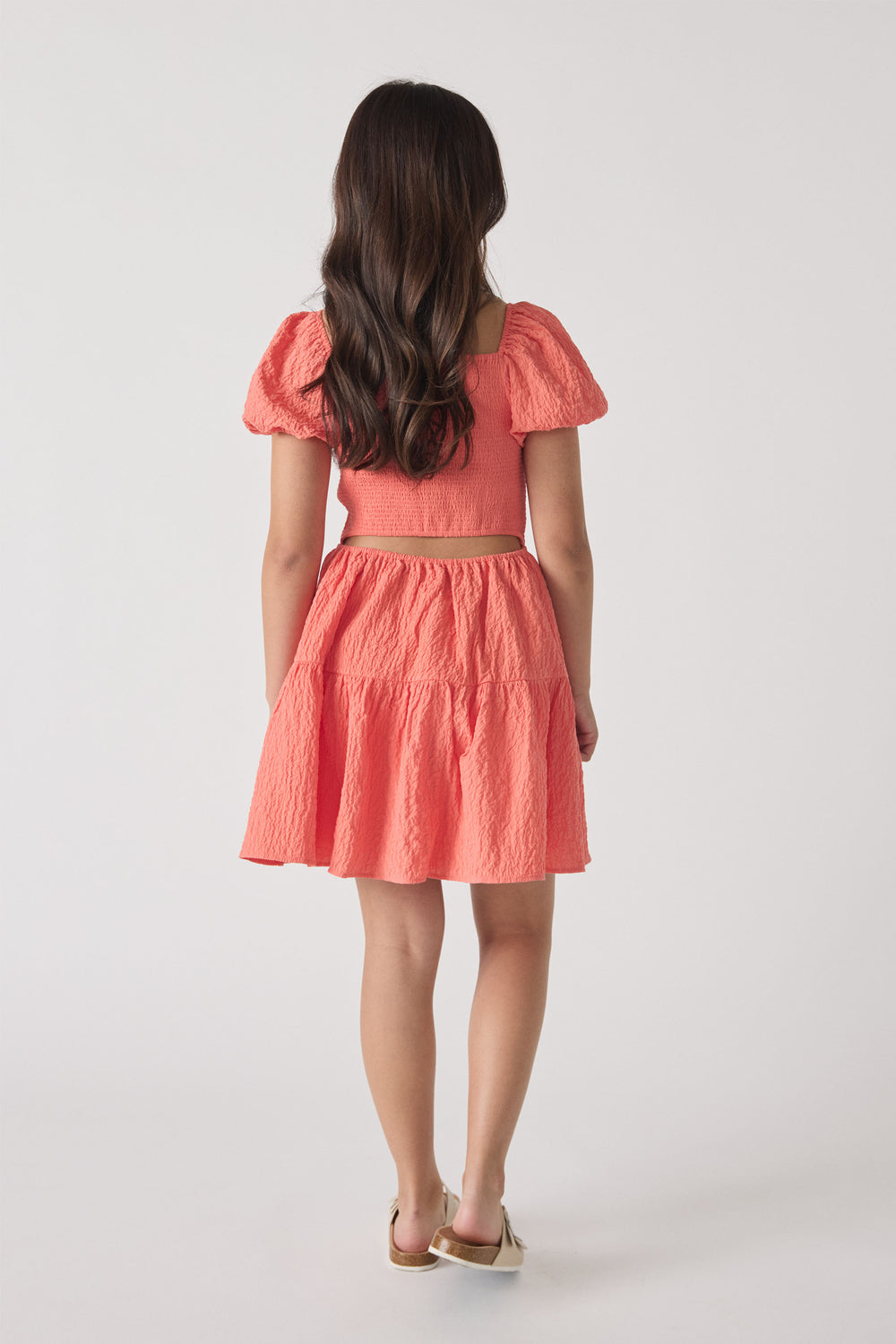 Girl wearing a textured coral mini dress with puffed sleeves and a tiered skirt, shown from the back.