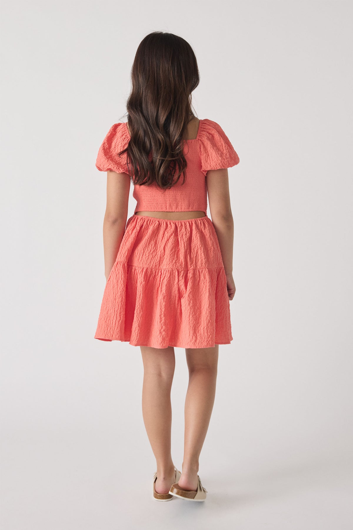 Girl wearing a textured coral mini dress with puffed sleeves and a tiered skirt, shown from the back.
