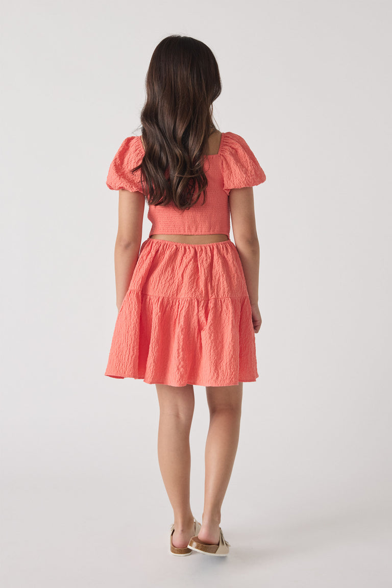 Girl wearing a textured coral mini dress with puffed sleeves and a tiered skirt, shown from the back.