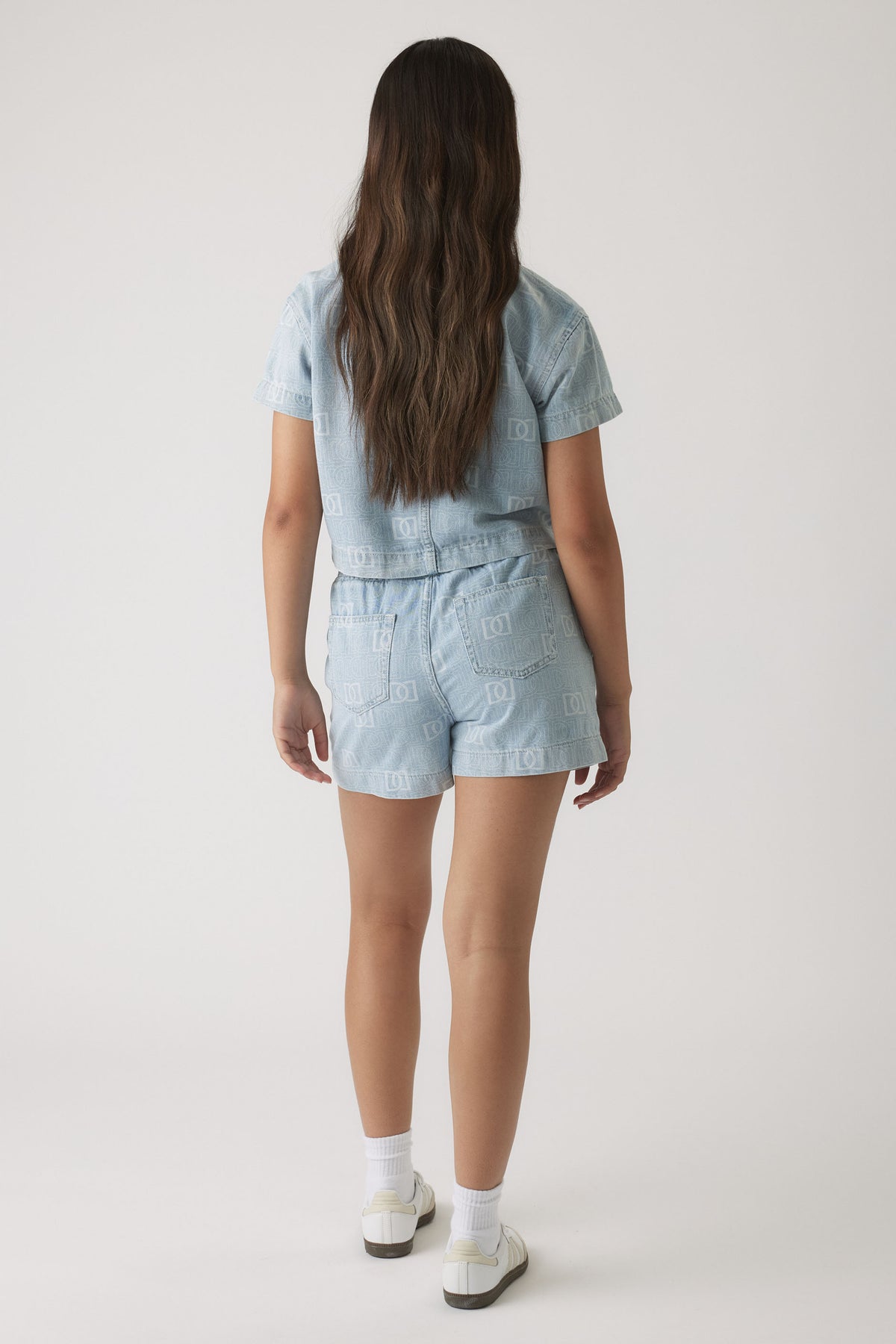 Girl wearing light wash denim shorts and matching short-sleeve jacket with subtle monogram pattern, styled with white sneakers.