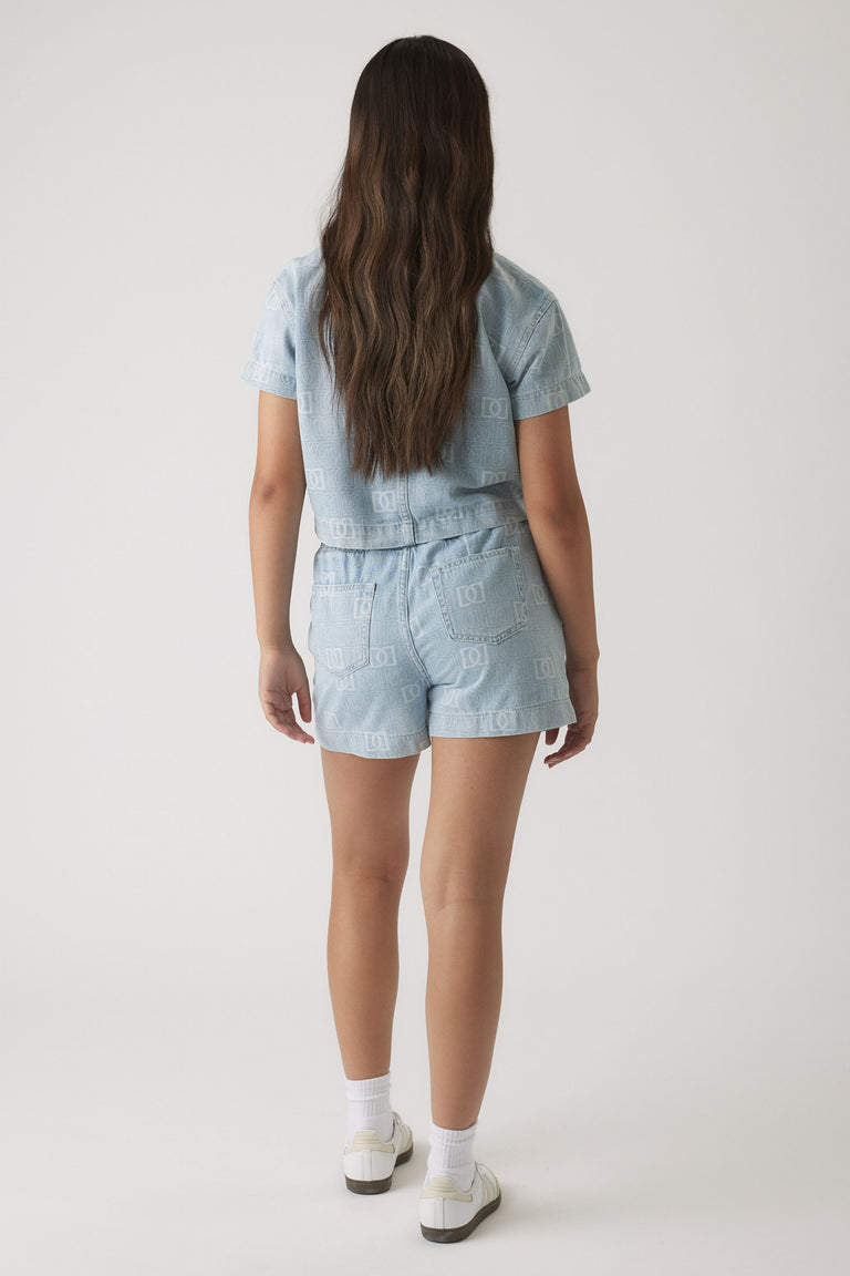 Girl wearing light wash denim shorts and matching short-sleeve jacket with subtle monogram pattern, styled with white sneakers.