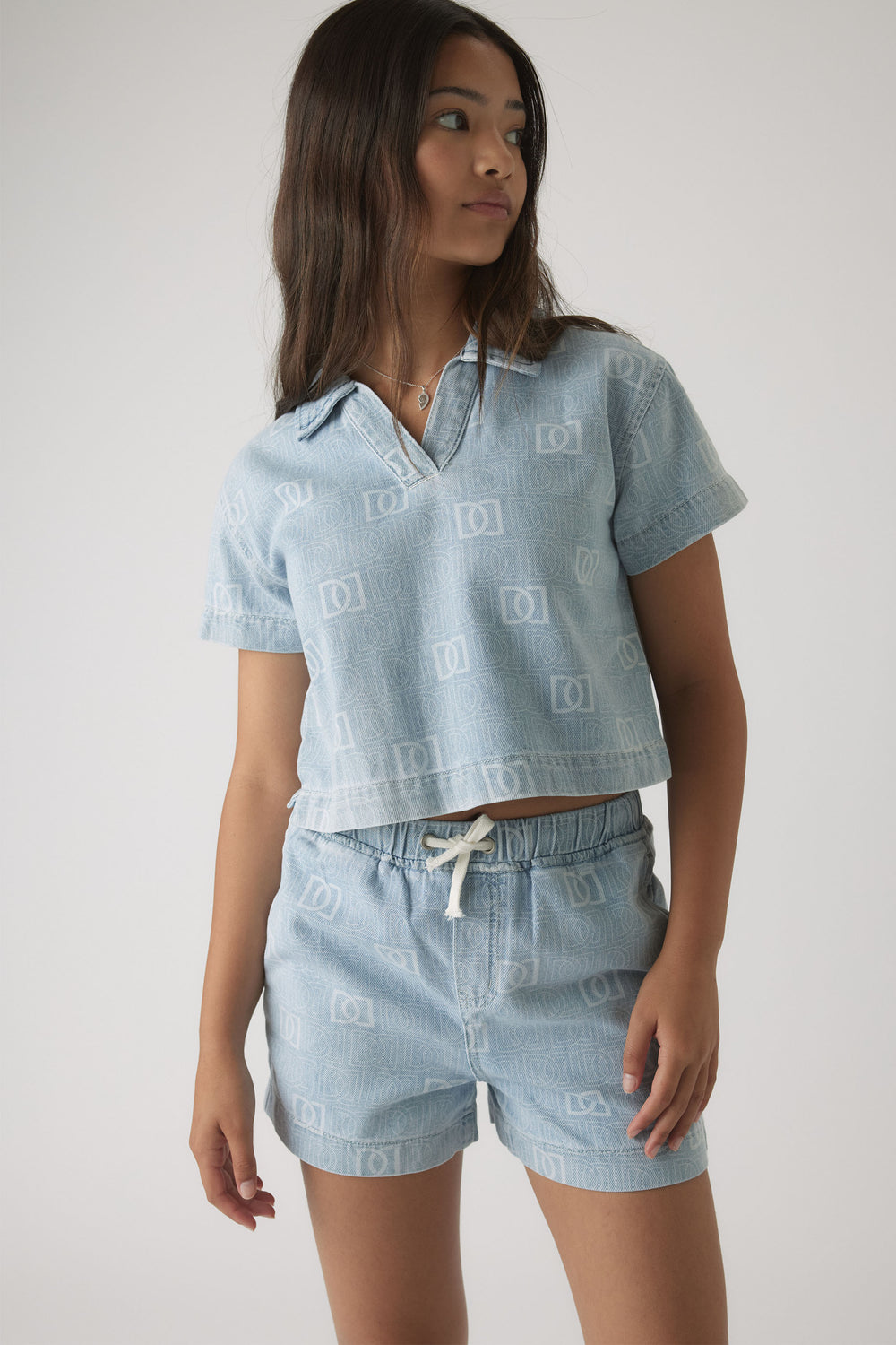 Girl wearing a light blue denim shirt and matching shorts set with subtle monogram print and relaxed fit.