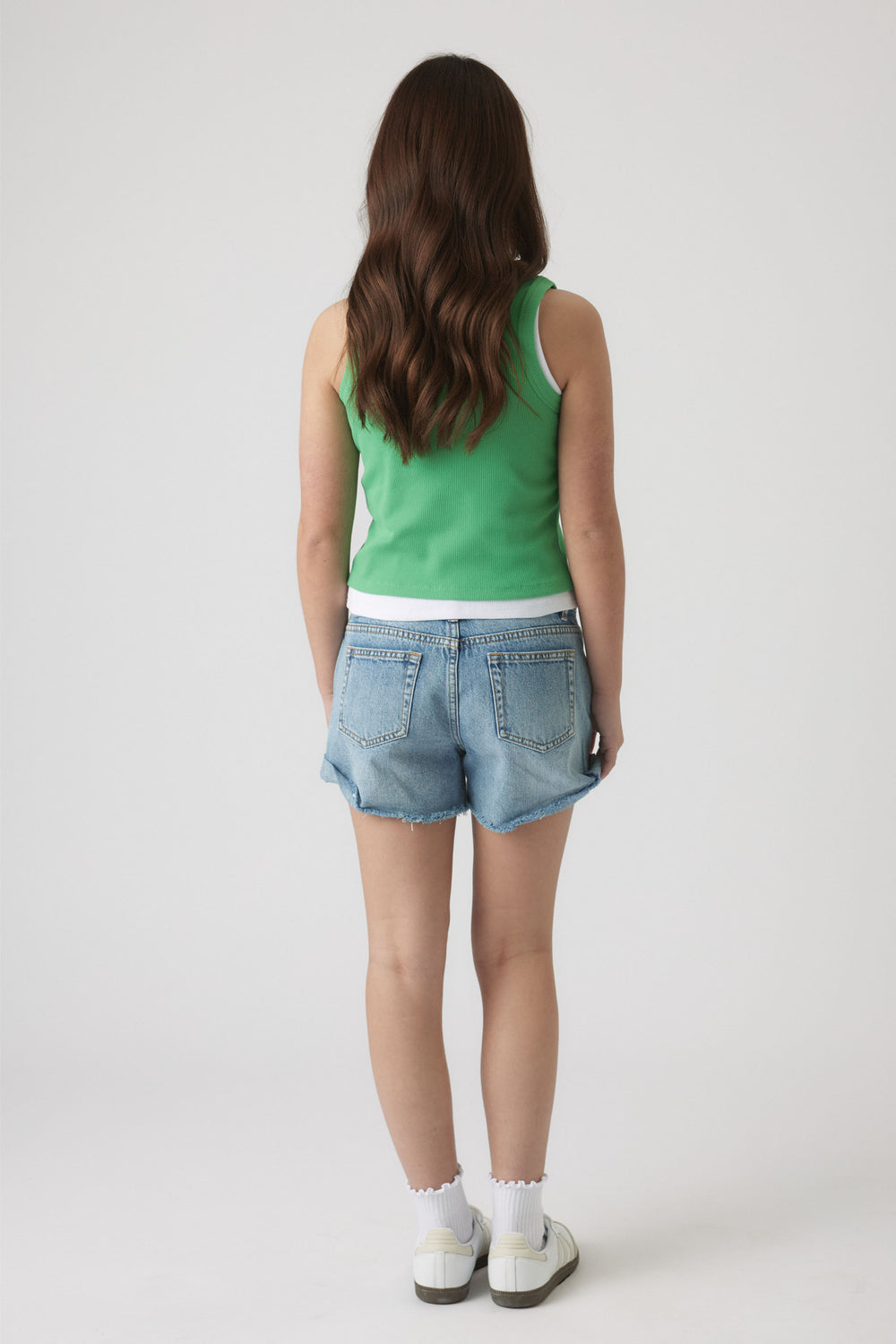 Girl wearing a green tank layered over a white tee paired with light blue denim shorts and white sneakers.