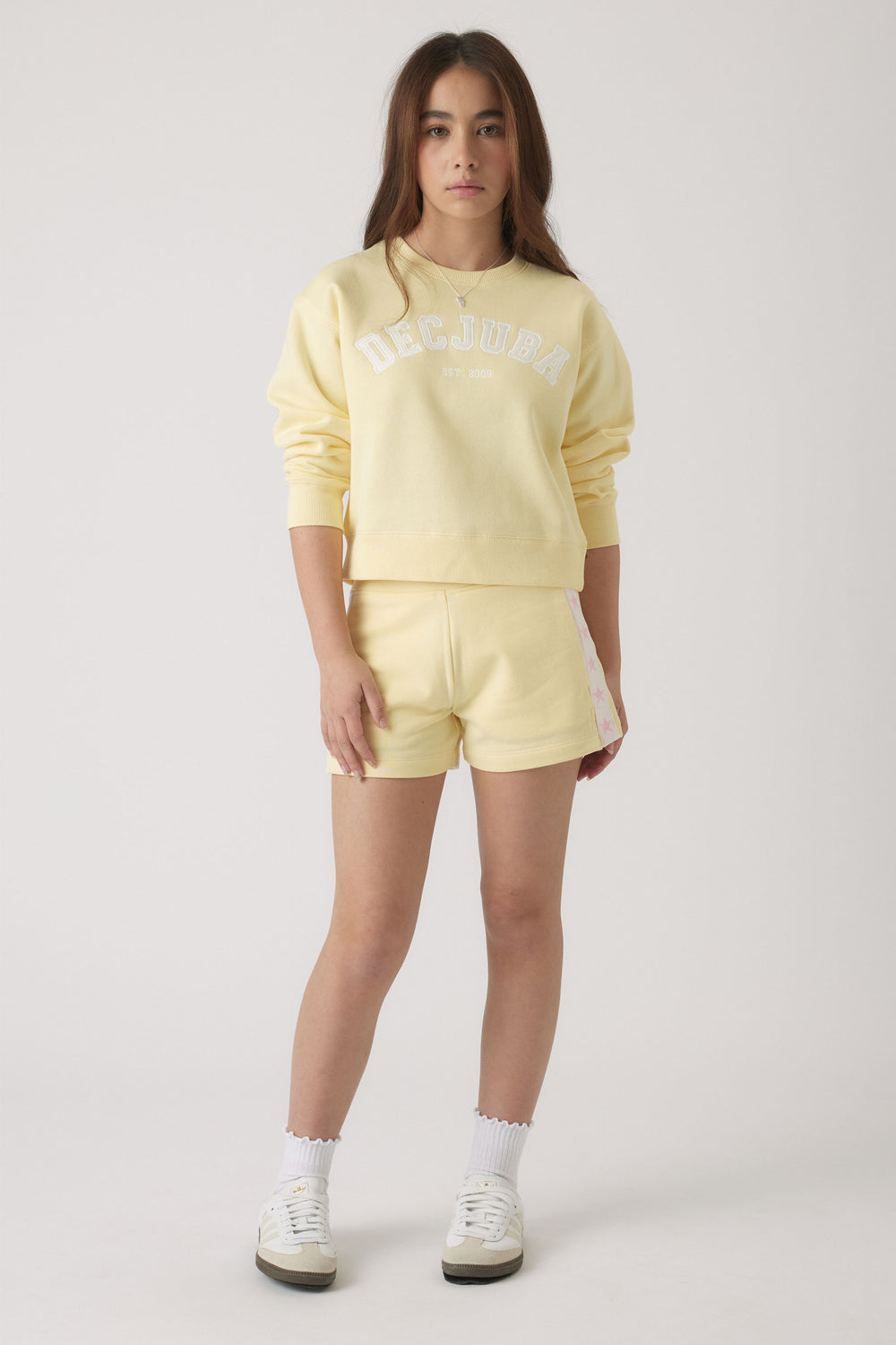 Teen girl wearing a soft yellow logo sweatshirt paired with matching shorts and white sneakers, standing front-facing.