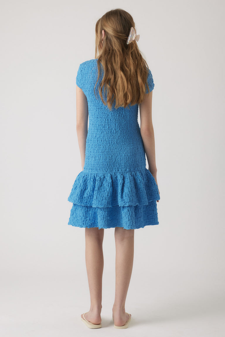 Girl wearing a sky blue shirred dress with short sleeves and a tiered ruffle hem, styled with a hair clip and slippers.