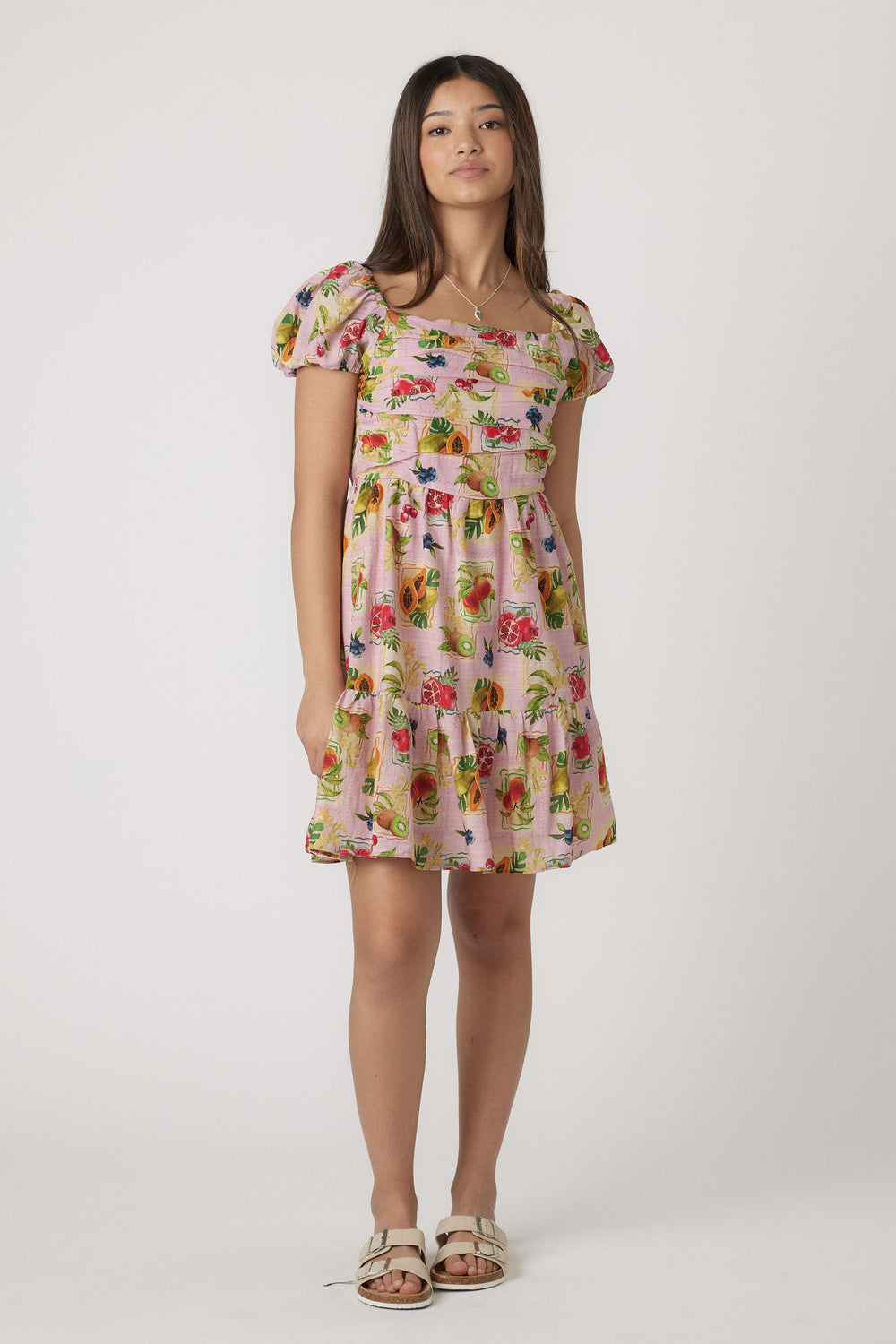 Girl wearing a pink mini dress with puff sleeves and colorful fruit print paired with cream sandals in a simple studio setting